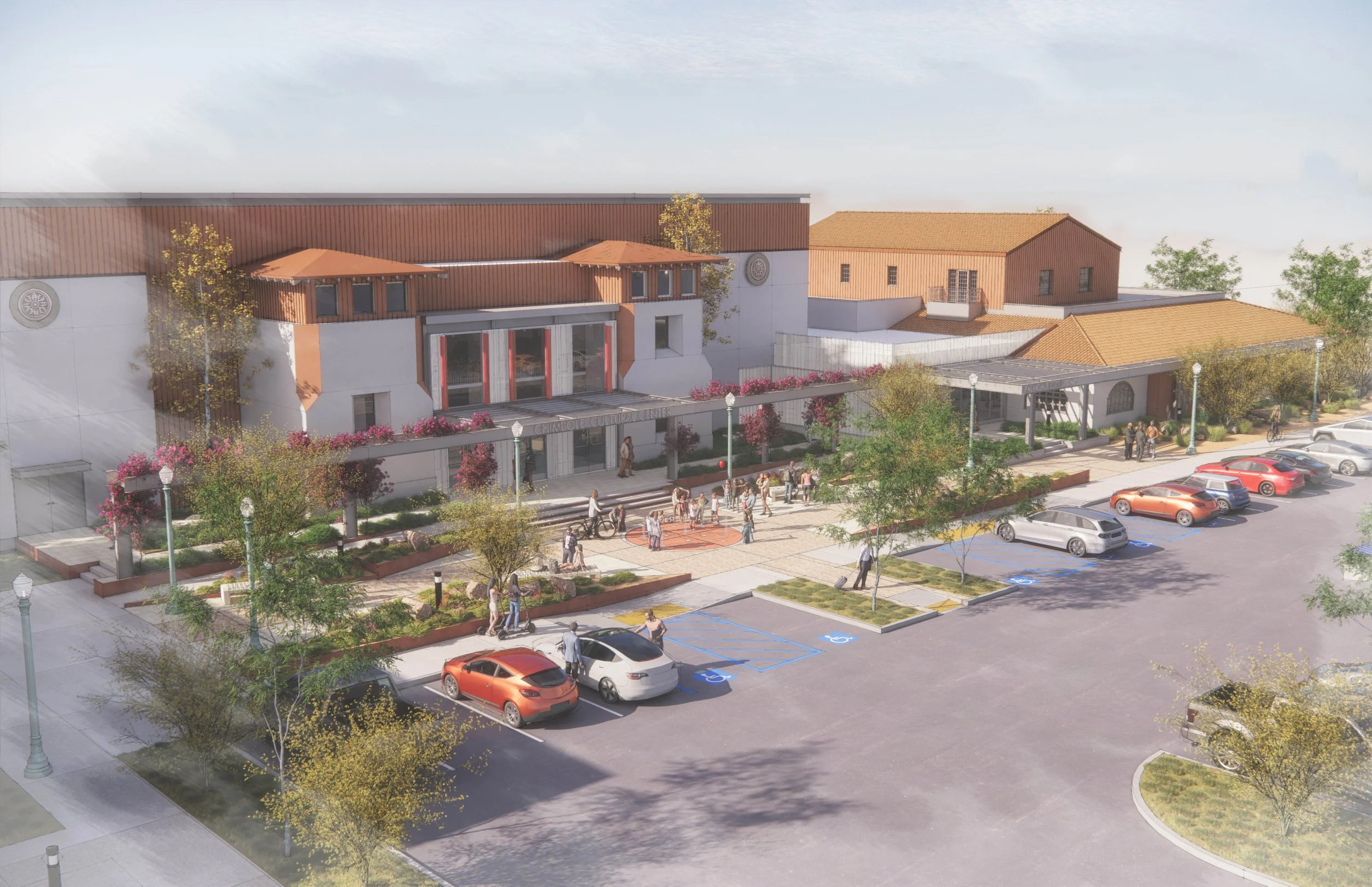 A modern community center with a parking lot, including designated handicap spaces, surrounded by trees and landscaping. People are walking and gathering outside the building, which features a white facade with red accents and outdoor seating.