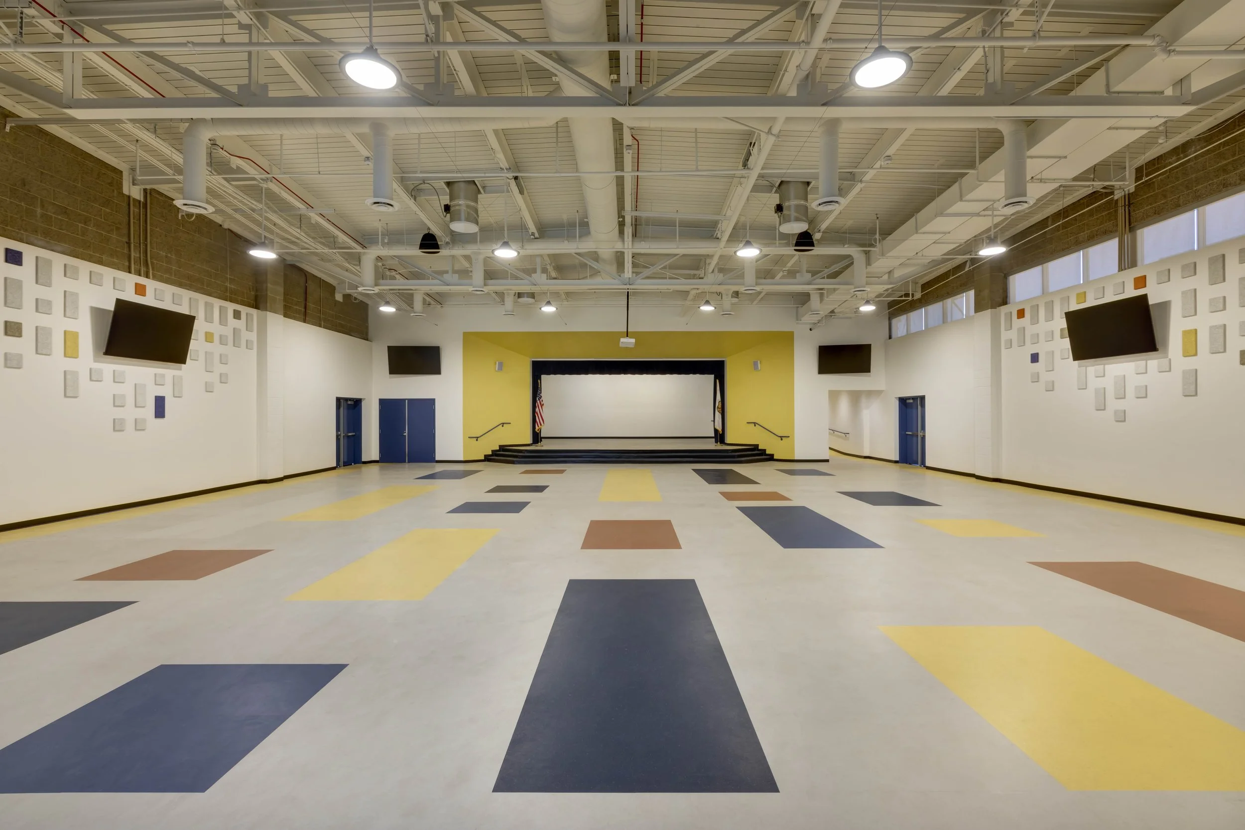 Empty school auditorium with a stage, American flags, four large wall-mounted televisions, and a colorful geometric patterned floor.