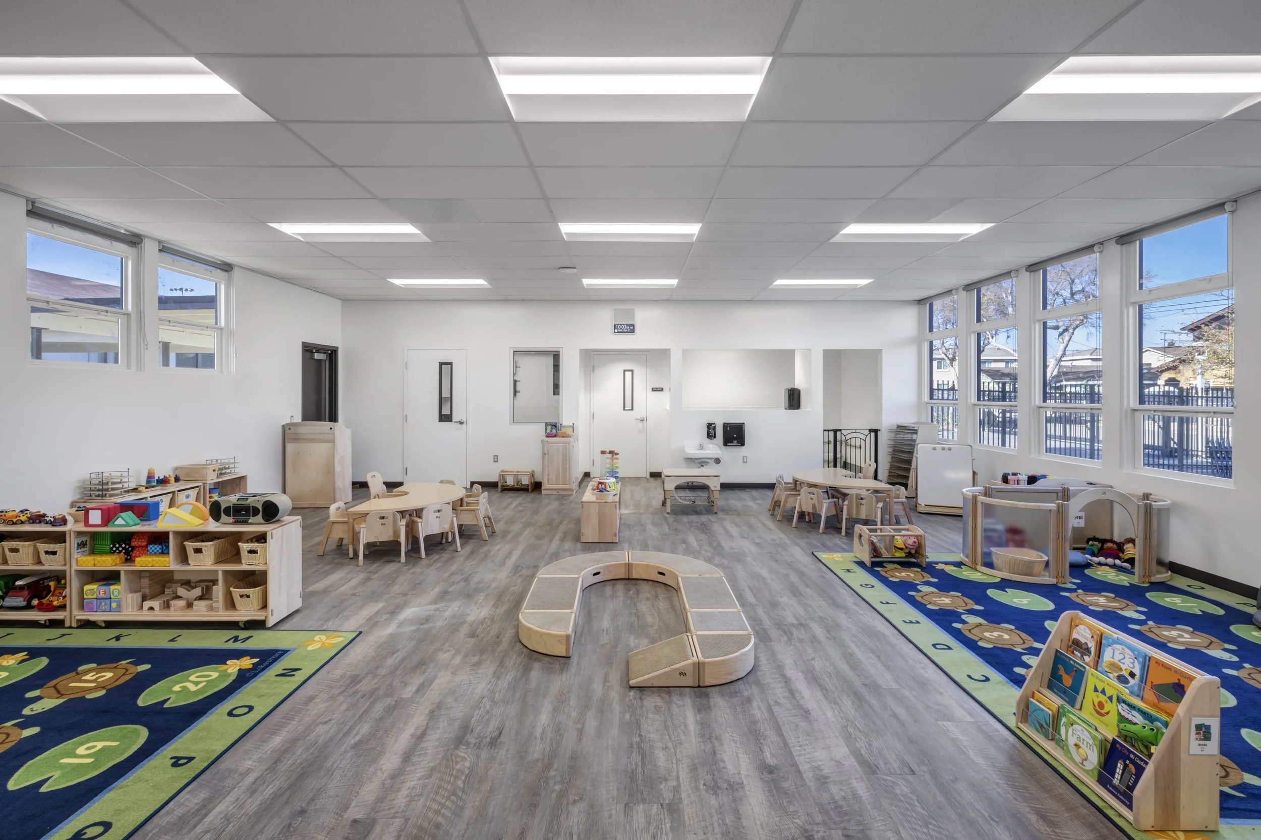 Bright and spacious early childhood classroom with large windows, colorful rugs, wooden tables and chairs, and various toys and educational materials.