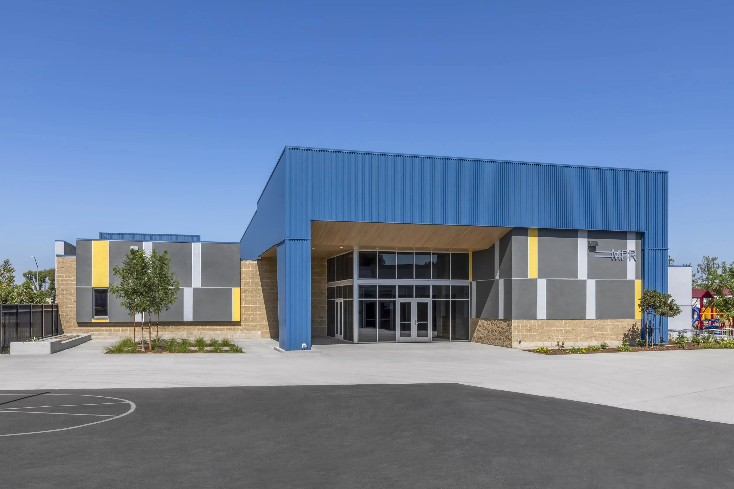 Modern school building with colorful exterior, large glass entrance, adjacent playground, and parking lot under a clear blue sky.