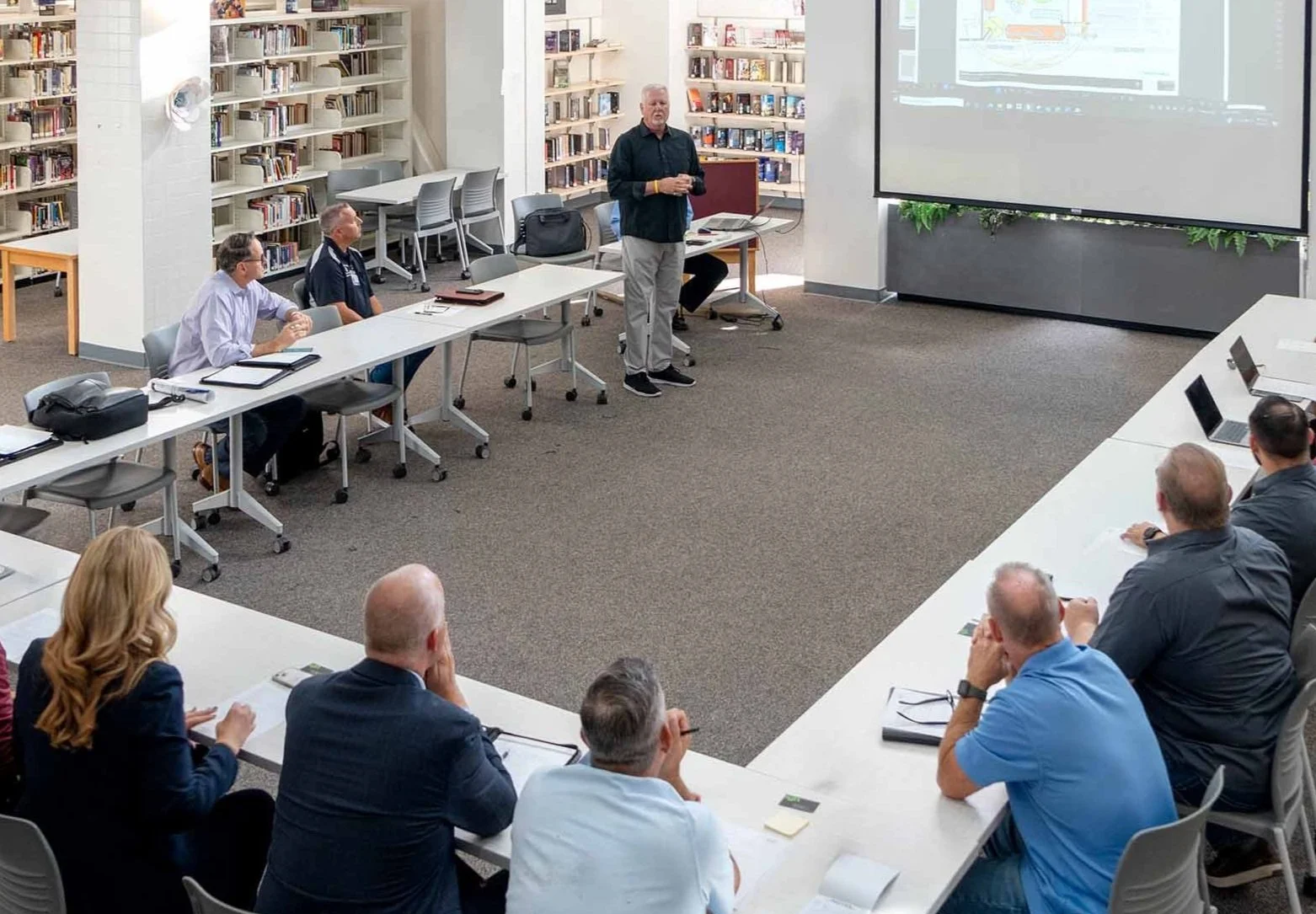 A man giving a presentation to a group of professionals in a library conference room. The audience is seated around a U-shaped table, facing the presenter and a large screen displaying a presentation slide.