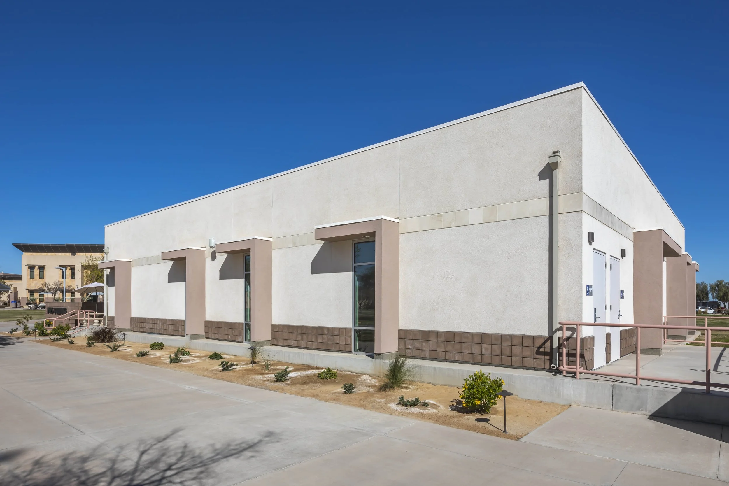 A modern, white commercial building with pink accents and a brown brick base, under a clear blue sky, with a paved parking area and small landscaped patches in the foreground.
