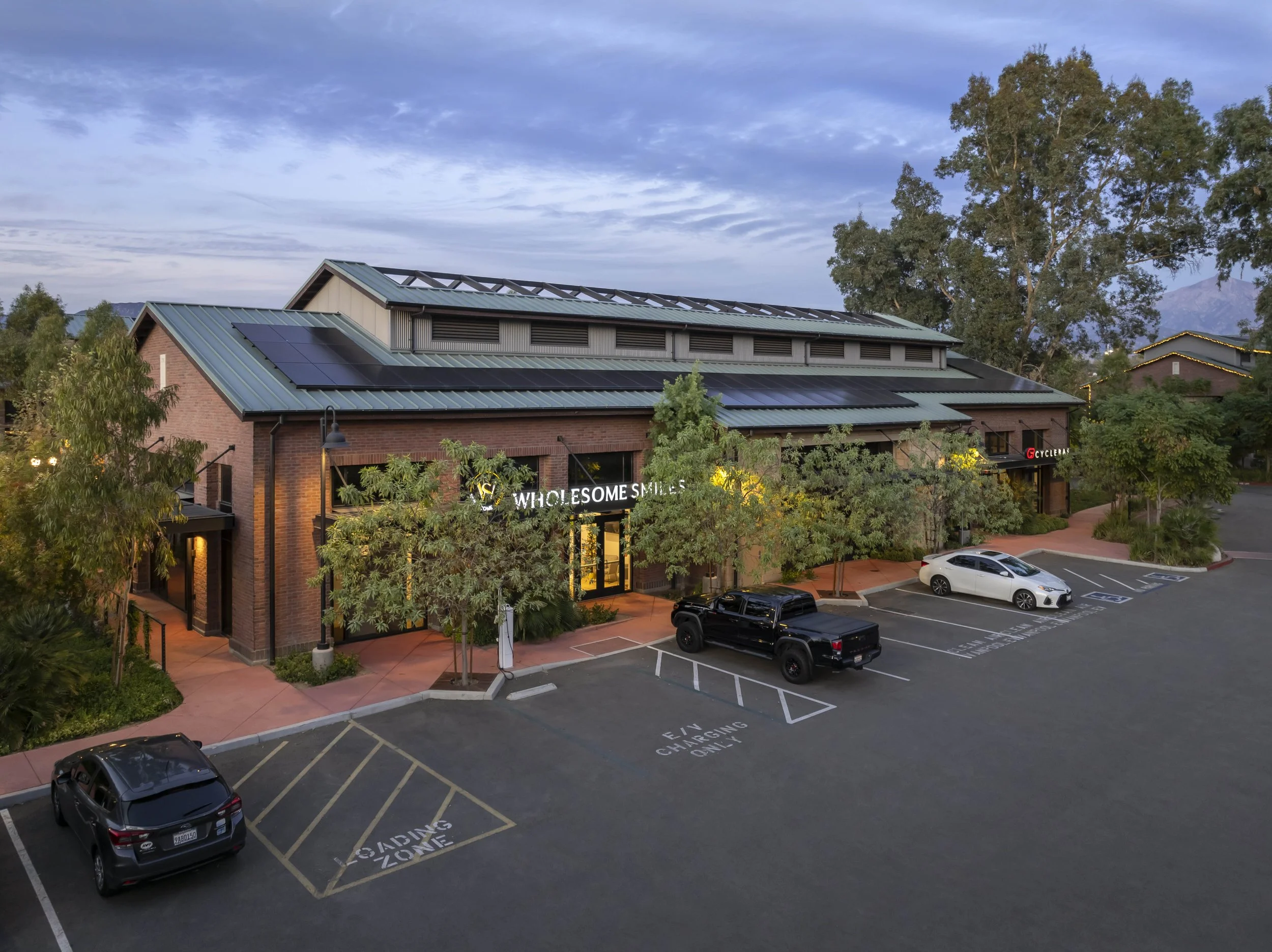 A strip mall with brick exterior walls and solar panels on the roof, surrounded by trees and parking lots, during dusk.