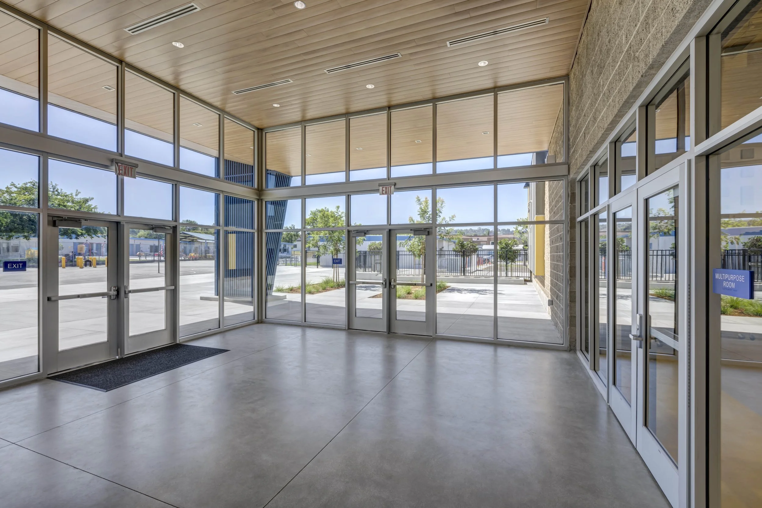 Empty modern building entrance with large glass windows and doors, a wooden ceiling, concrete floor, and outside view of trees and parking lot.