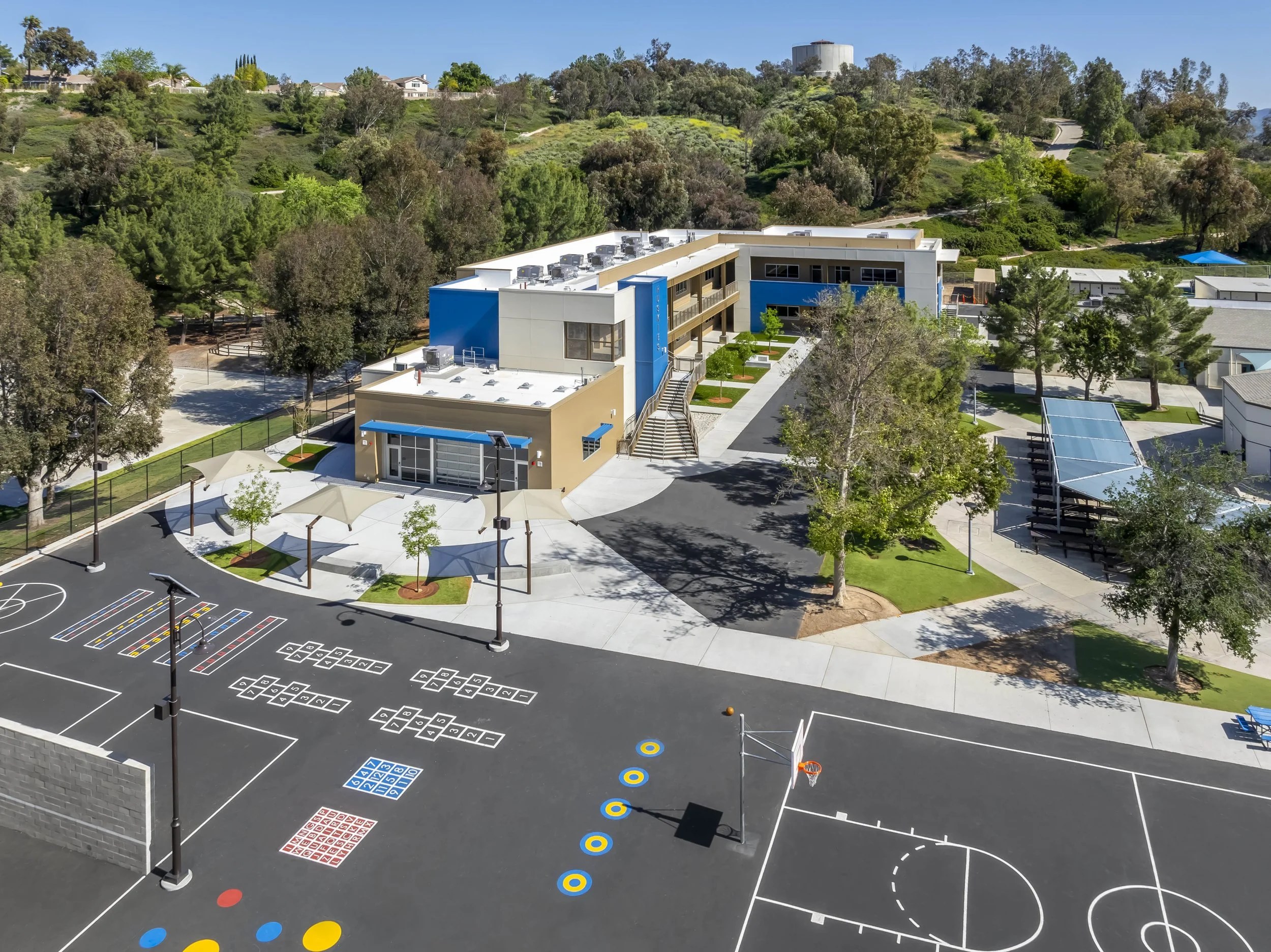 A modern school building with a playground featuring multiple basketball courts, hopscotch, and hop balls, surrounded by trees and green landscaping, under a clear blue sky.