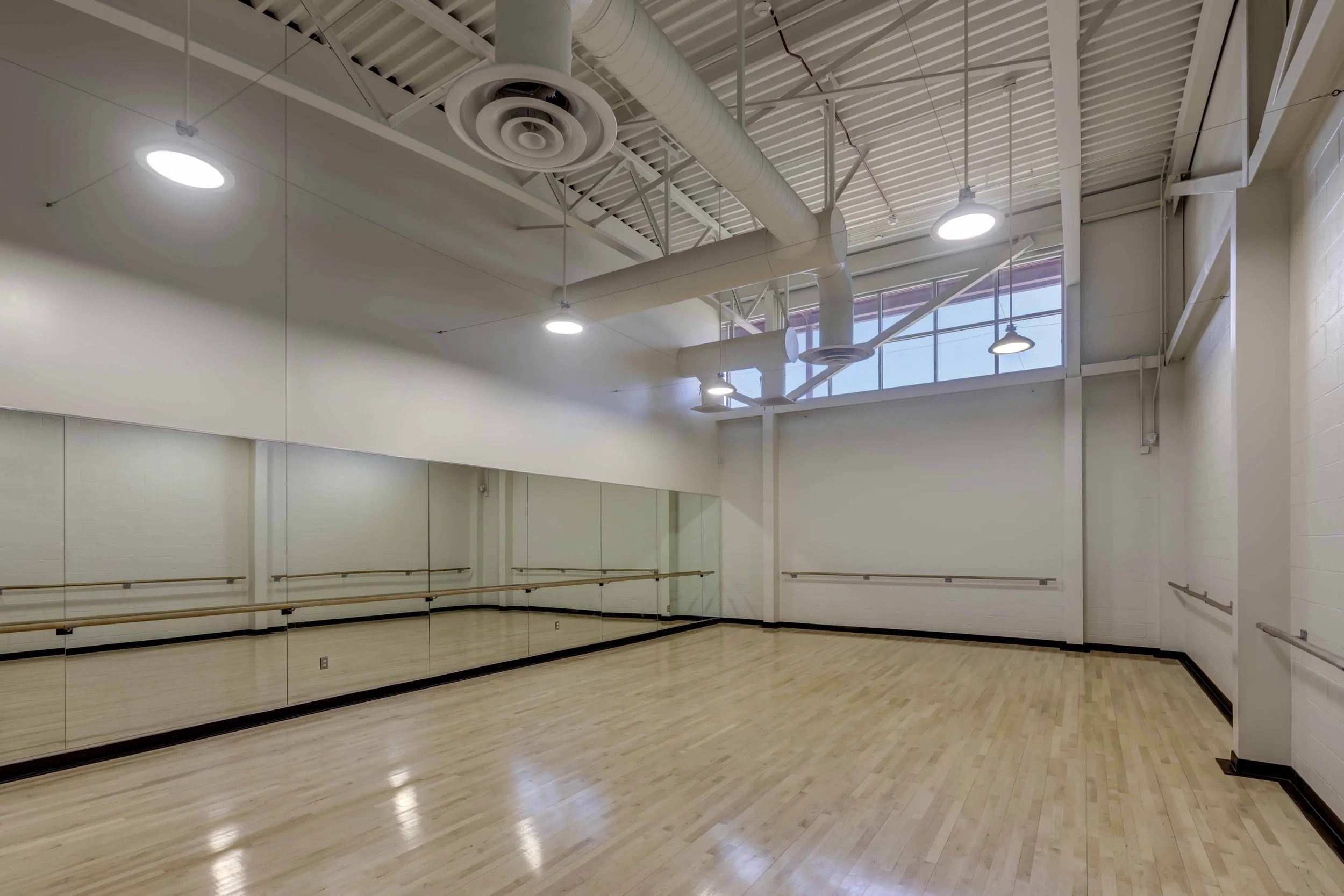Empty indoor dance or exercise studio with wooden floor, white walls, mirror wall, handrails, and ceiling lights.