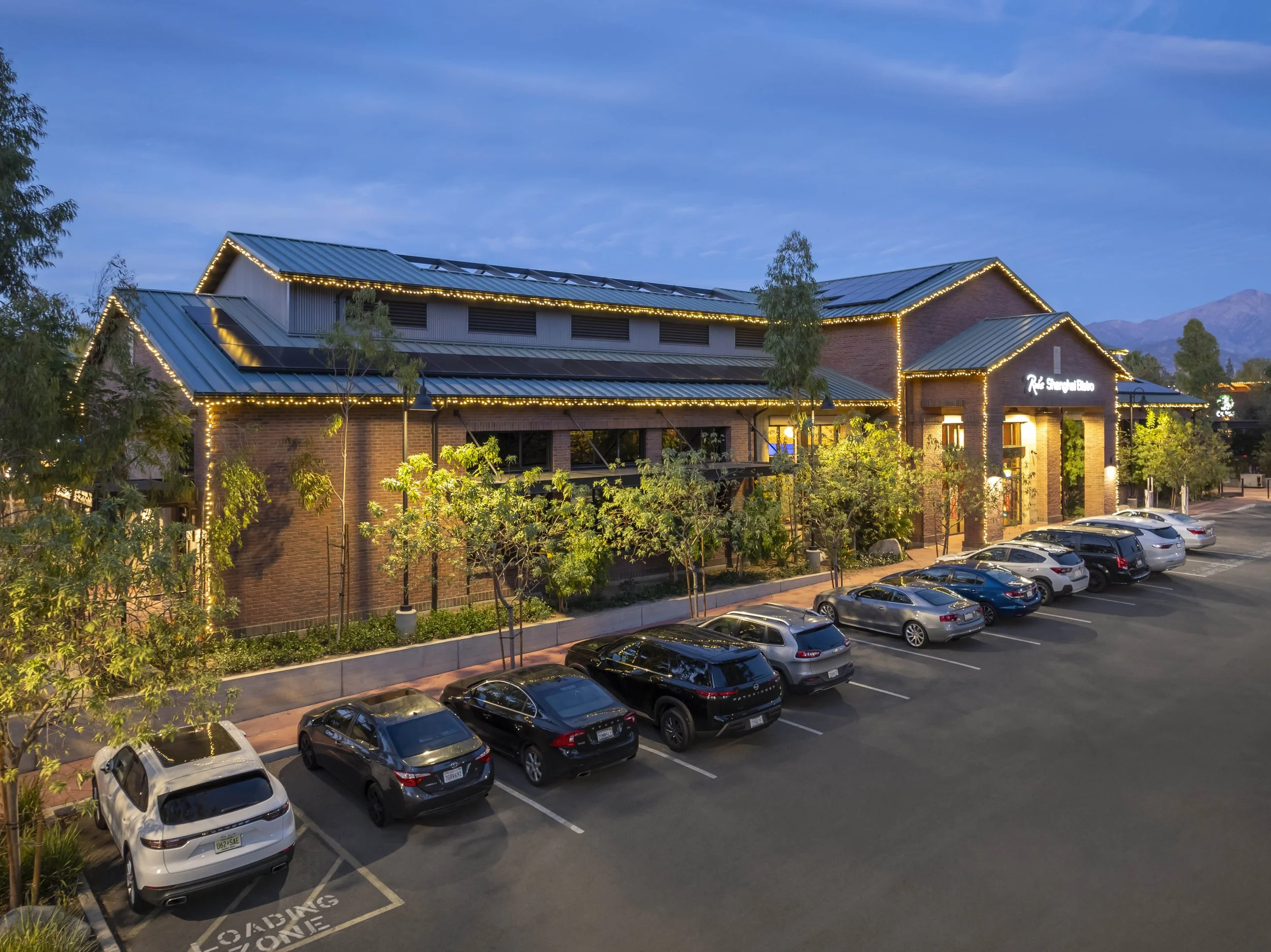 A commercial building decorated with string lights along the roofline at dusk, with a parking lot filled with cars in front and trees lining the sidewalk.
