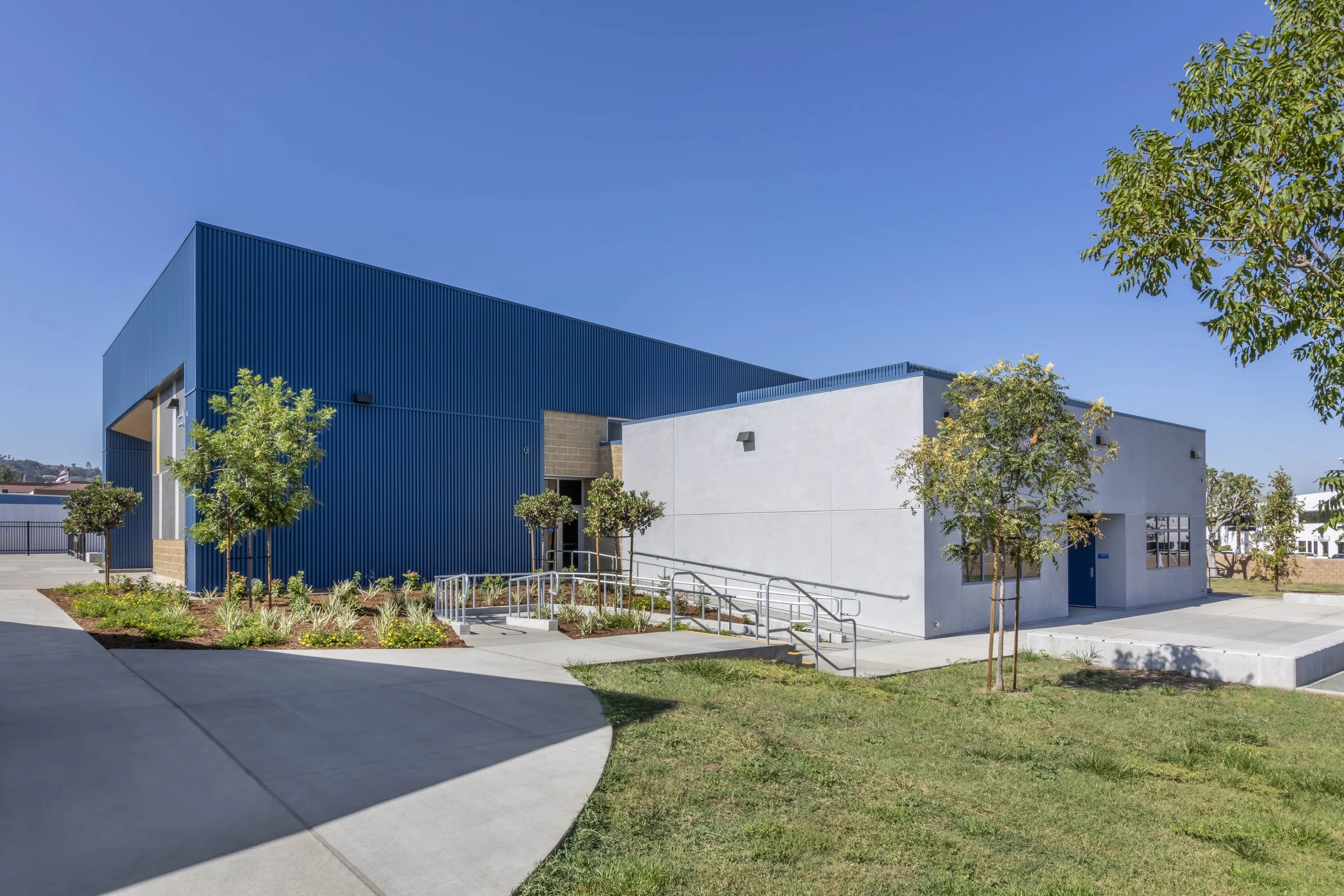 Modern building with blue and white exterior walls, surrounded by trees and landscaped garden, under a clear blue sky.
