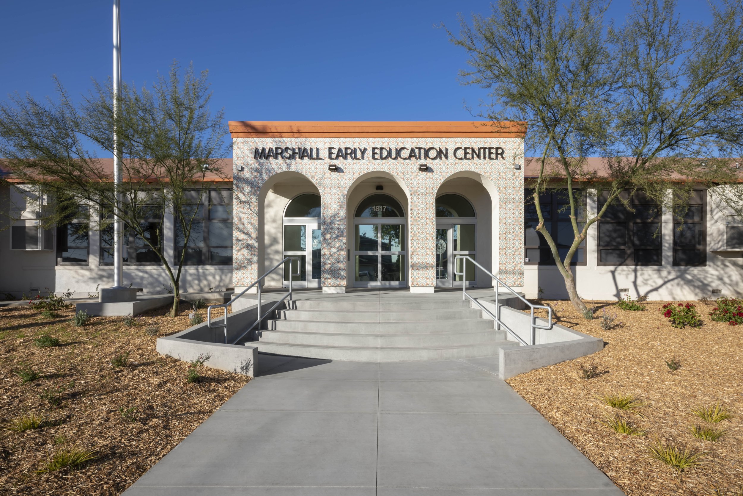Front view of Marshall Early Education Center building with stairs and ramp, surrounded by desert landscaping and trees, under a clear blue sky.