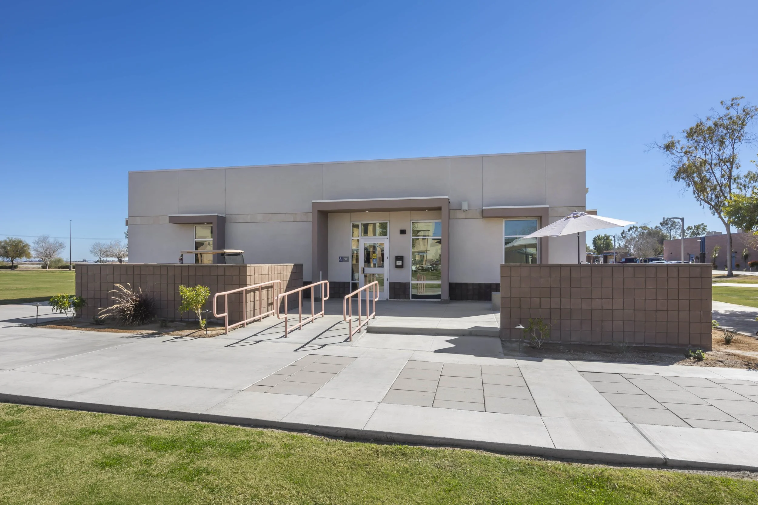 A modern, single-story building with a light-colored exterior and a small ramp in front, surrounded by a landscaped area with grass, plants, and trees. The building has glass doors and windows, with a white umbrella on the right side and a sidewalk l