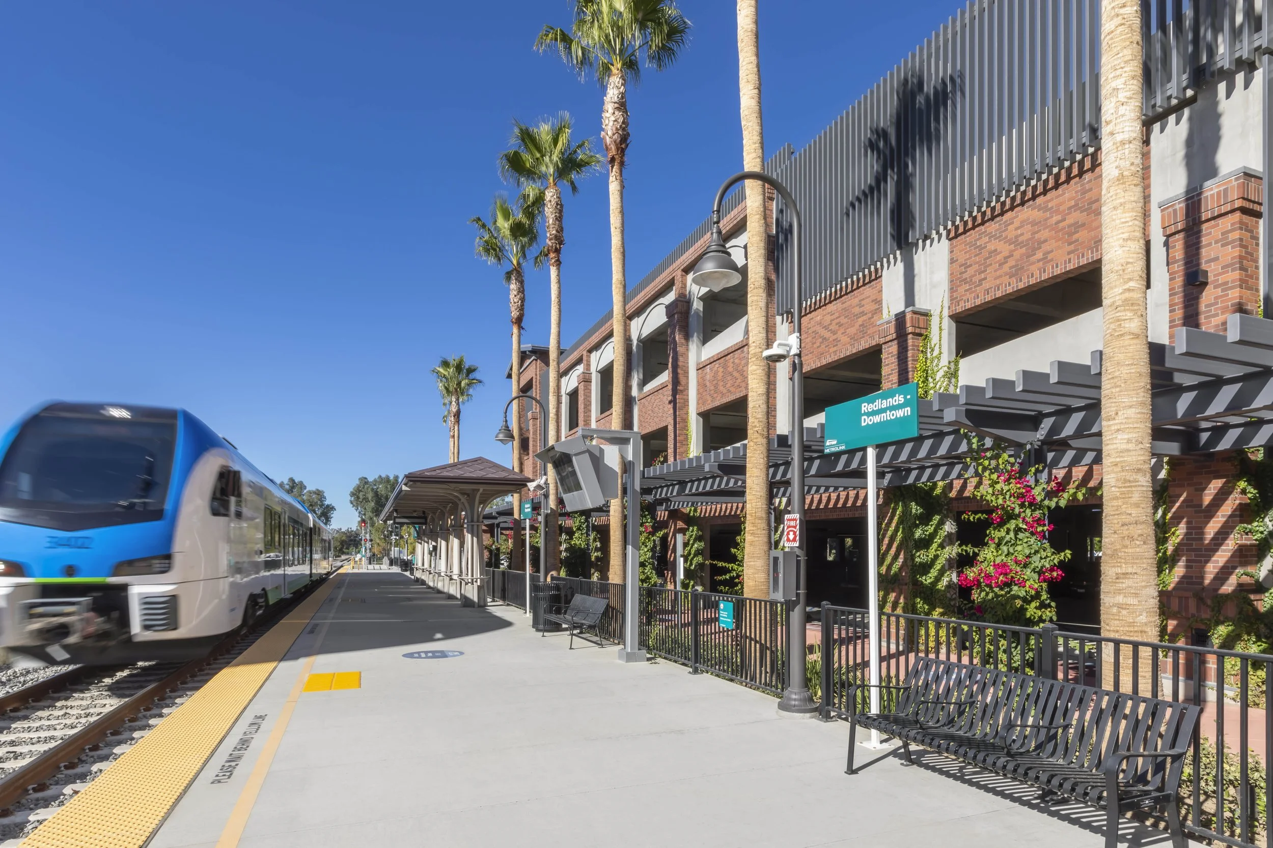 A train arriving at Redlands Downtown train station with palm trees, benches, and a brick building under a clear blue sky.