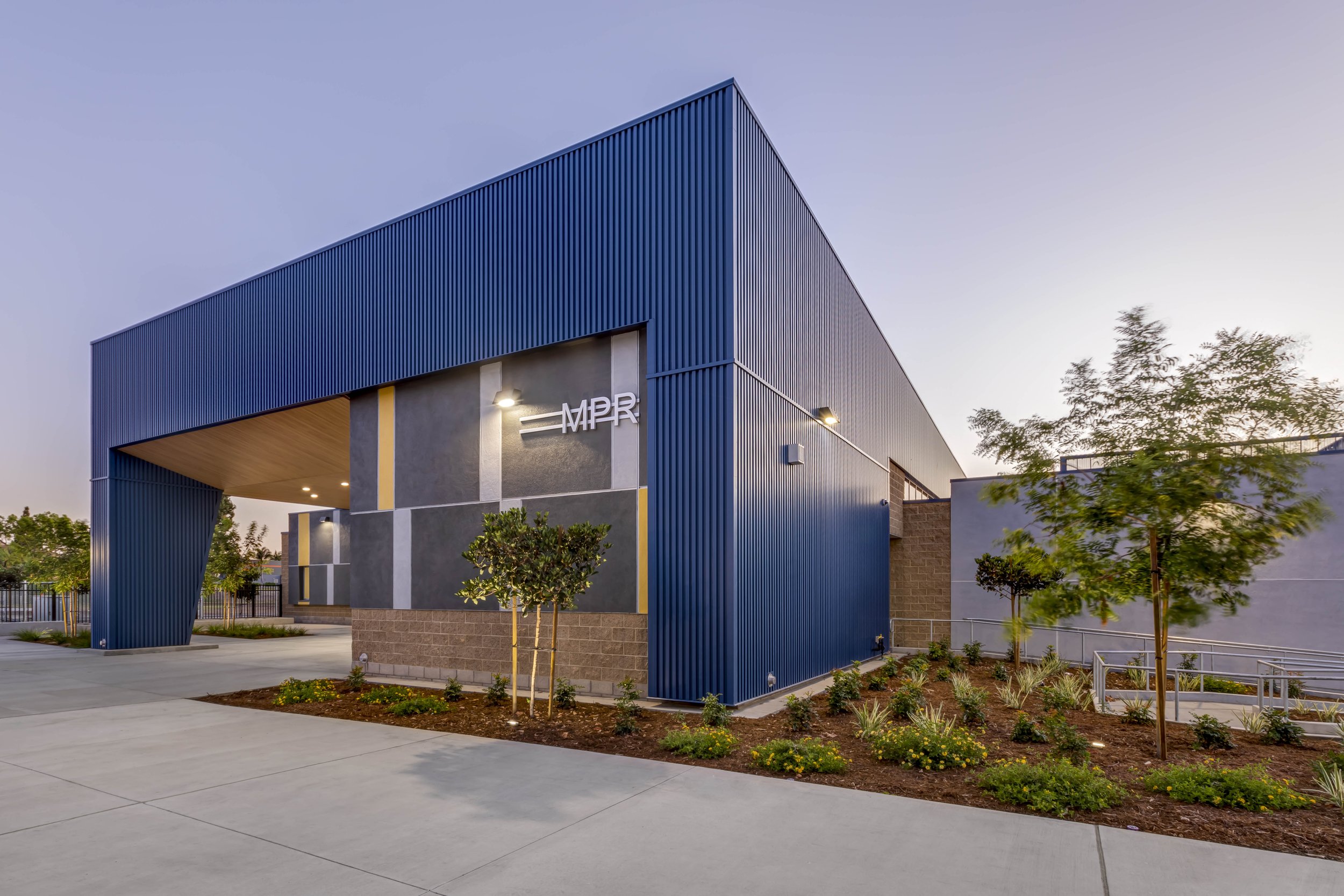Modern building with blue metal siding and logo 'MPR' on front, landscaped area with small trees and plants, sidewalk in foreground, clear sky in background.