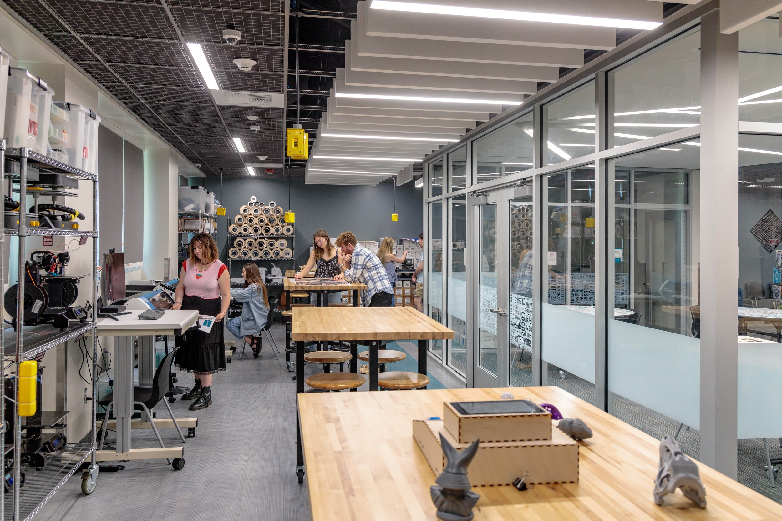 Interior of a modern workspace or workshop with people working at tables, shelves with equipment and materials, glass partition walls, and bright lighting.