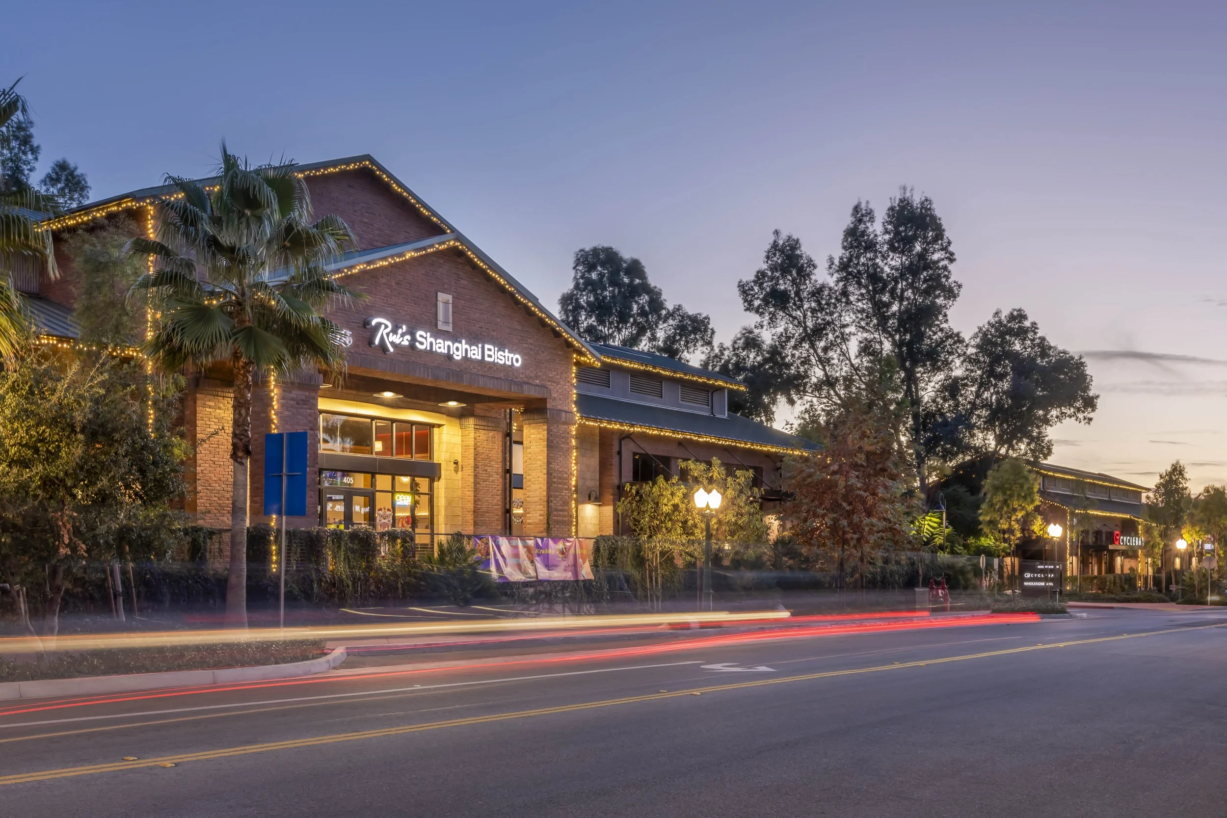 Exterior of Rio Shanghai Bistro at dusk with string lights, palm trees, and trees lining the street.