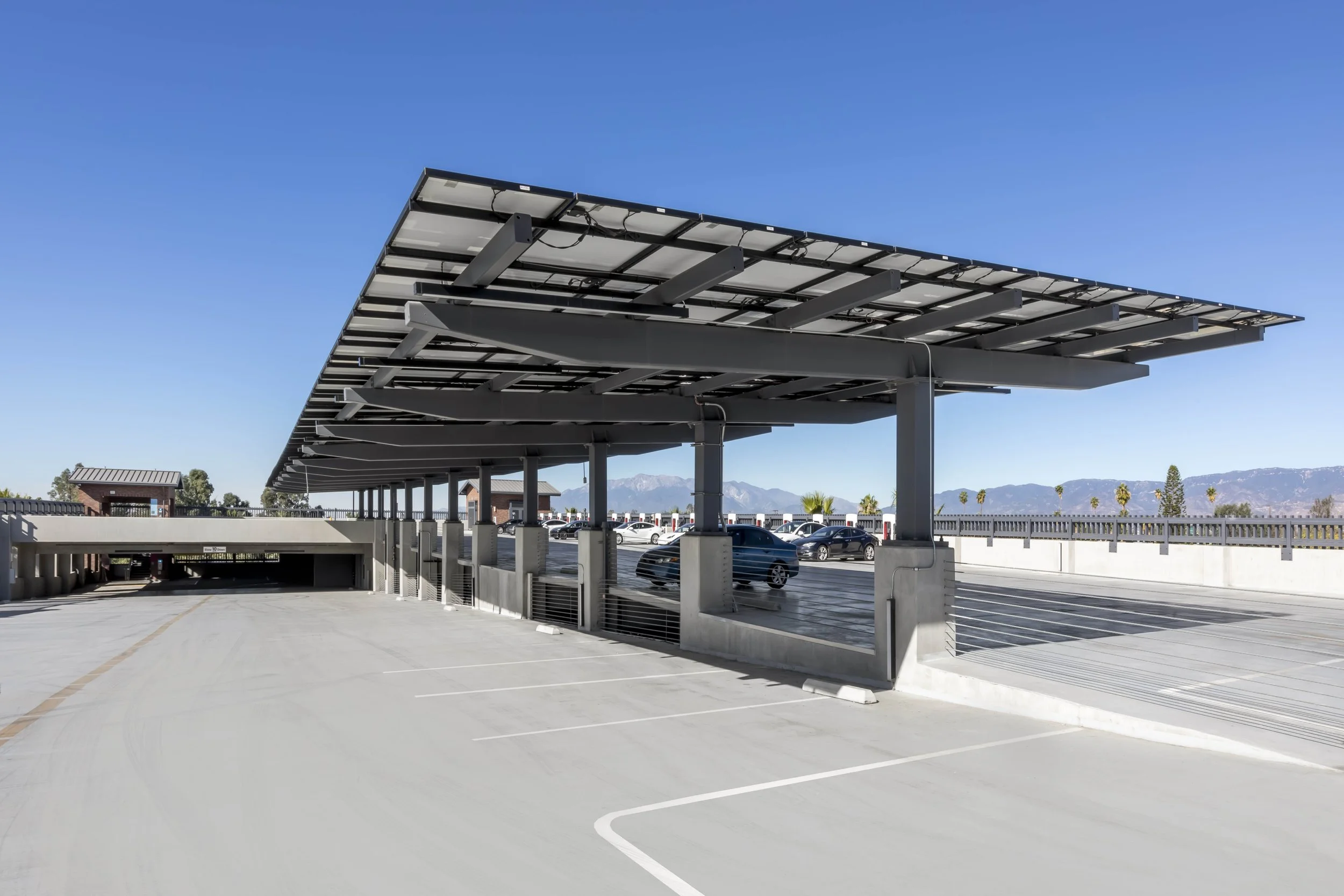 Parking lot with an electric vehicle charging station and solar panel shade structure near an open parking area, with mountains in the background.