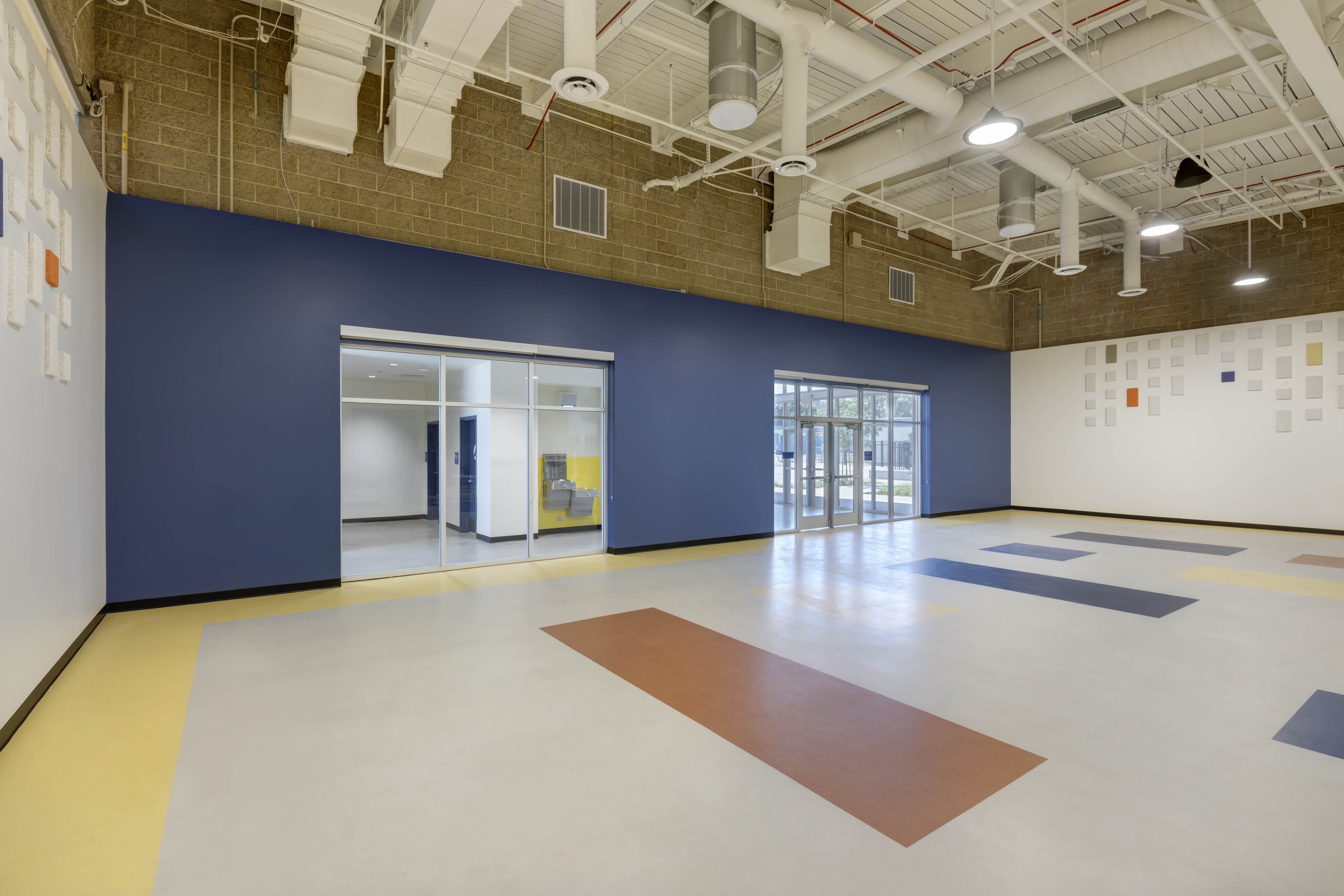 Empty indoor space with colorful tile flooring, blue accent wall, glass doors, and an industrial ceiling ceiling with exposed ducts and pipes.