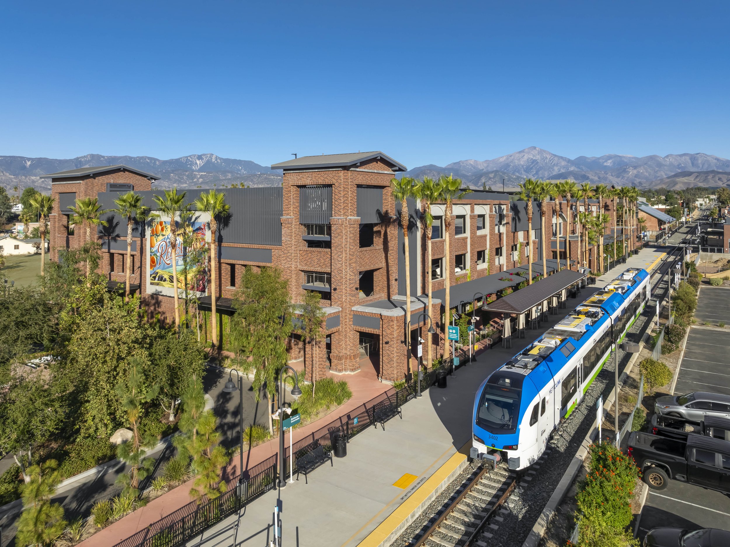 A modern train station with a blue and white train on the tracks, a brick building with outdoor patios, palm trees, parking lot, and mountains in the background.