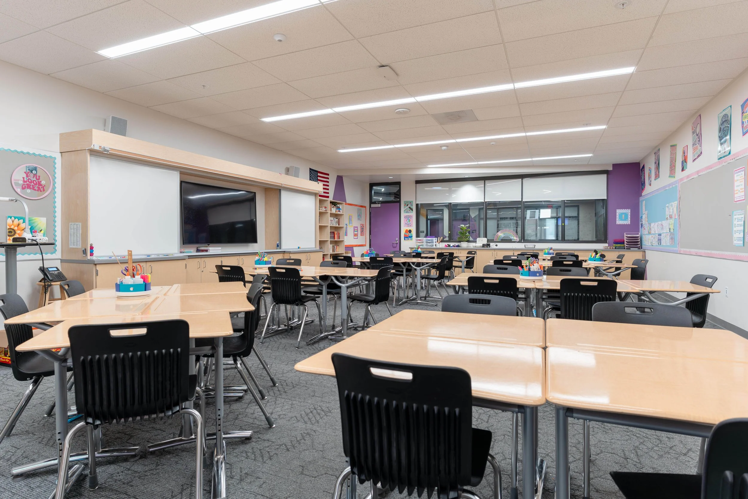 Empty classroom with multiple tables and chairs, whiteboard, large display screen, and colorful posters on the walls.
