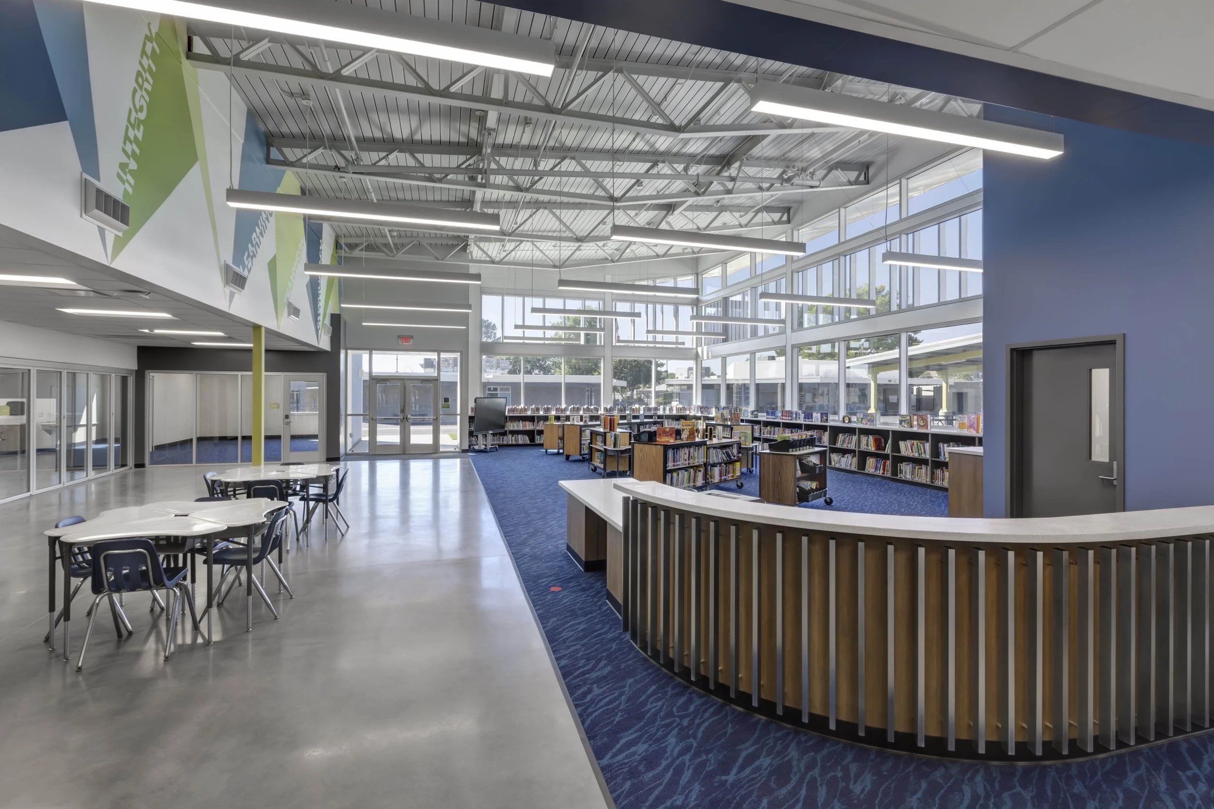 Interior of a modern school library with large windows, blue carpet, and bookshelves filled with books.