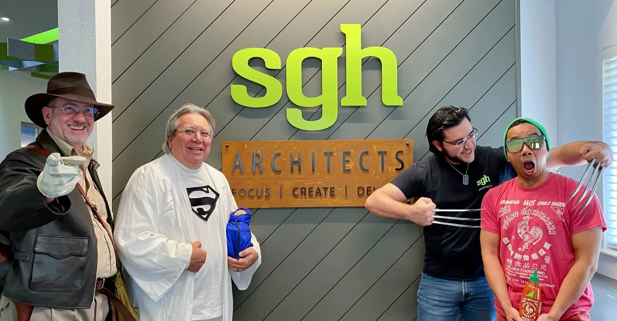 Four men posing playfully in front of a wall with the logo 'sgh ARCHITECTS'. One man dressed as Superman, one as Indiana Jones, one in casual clothing with glasses, and the last man wearing sunglasses and a T-shirt with chili sauce graphic, making a claw gesture with fake claws.