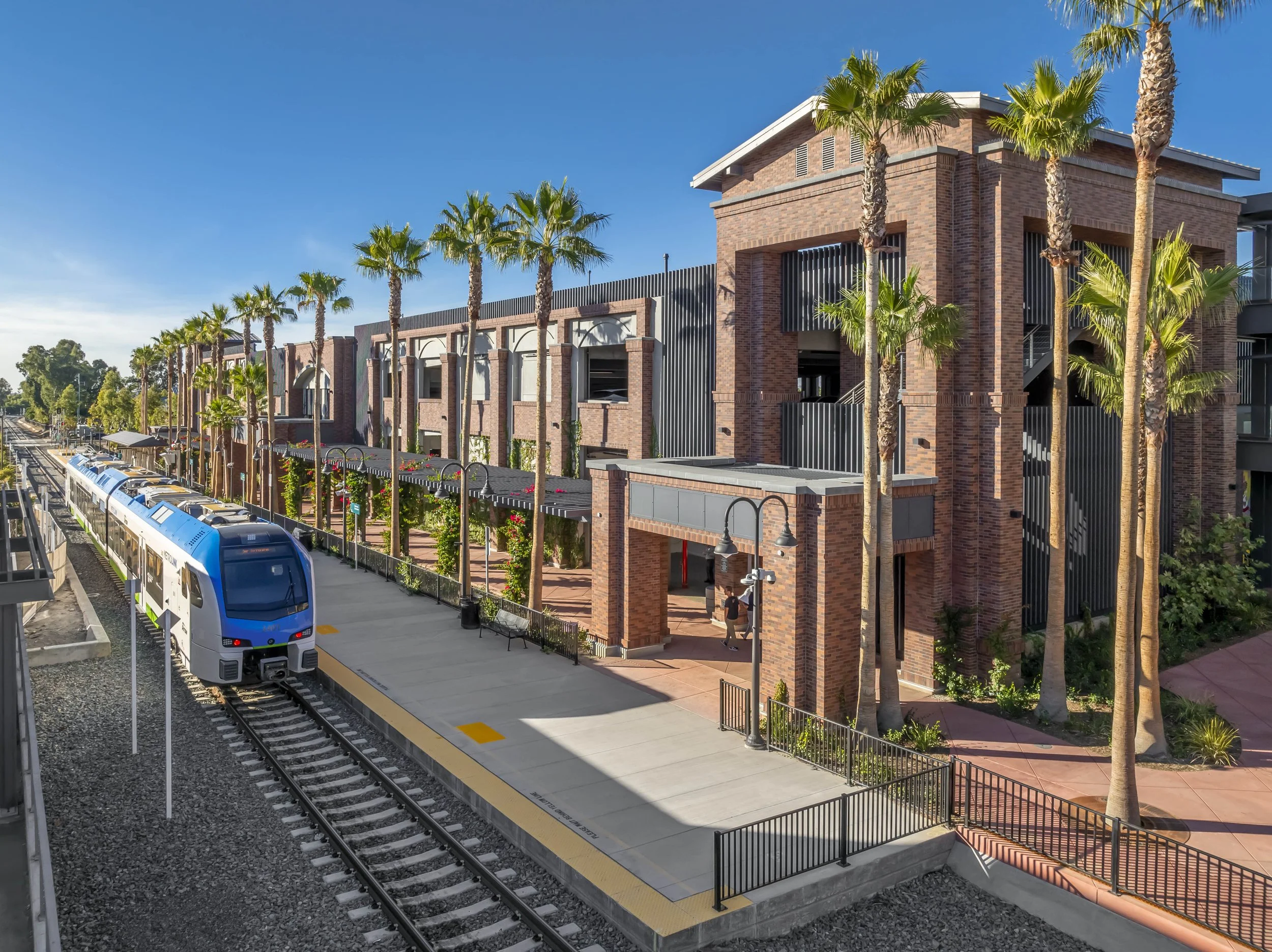 A modern brick building with palm trees lining a sidewalk next to a railway station. A blue train is stopped at the platform with a few people walking nearby, under clear blue skies.