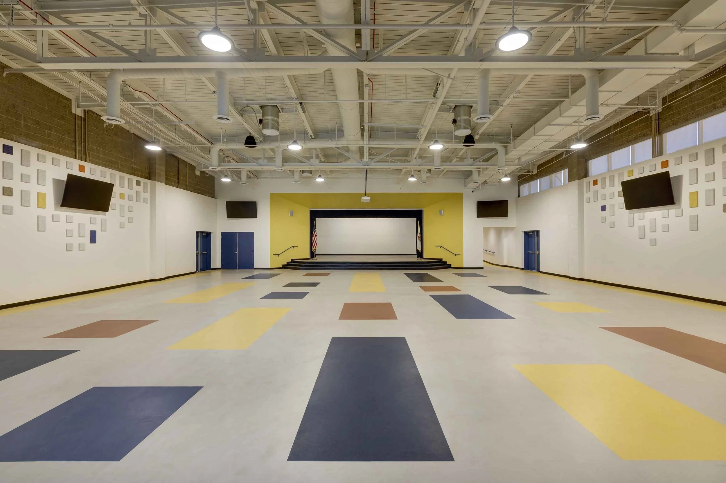 Empty multipurpose room with a stage, colorful floor tiles, yellow accent wall, and multiple wall-mounted screens.