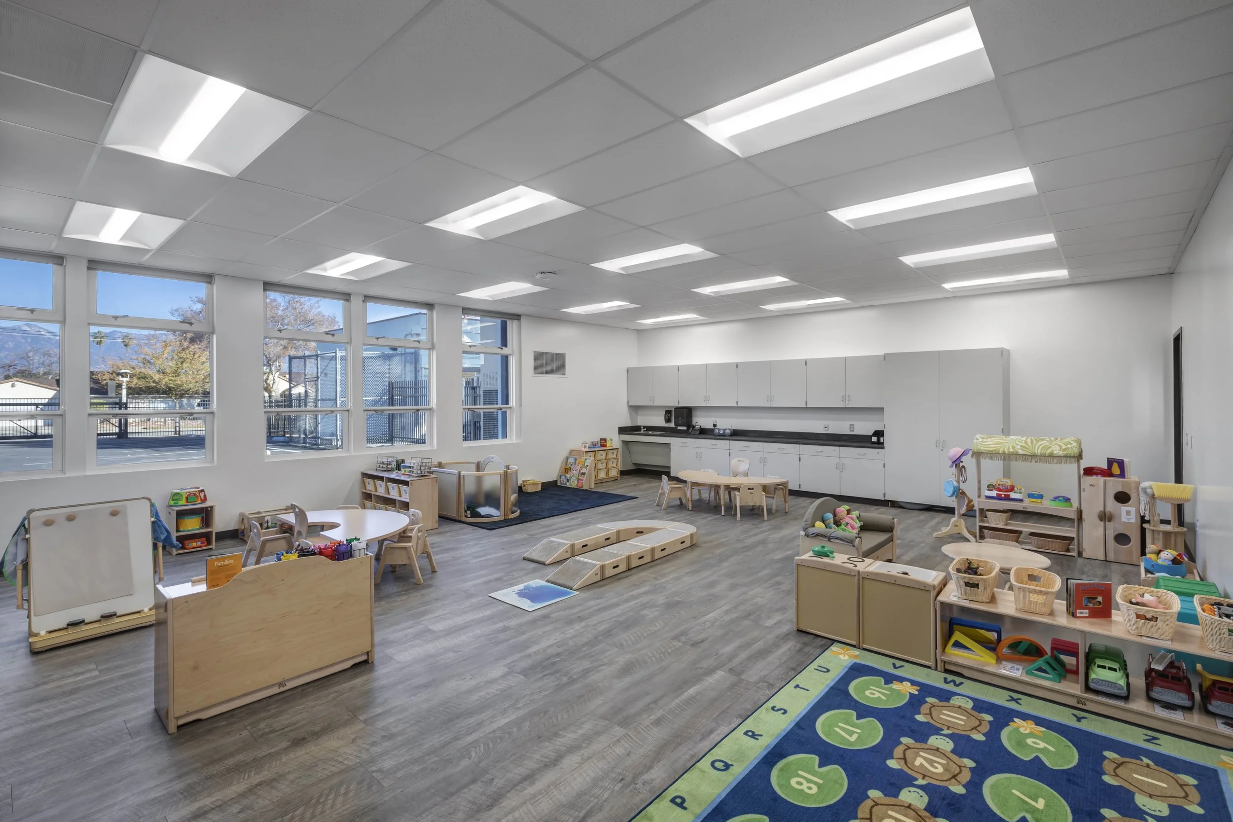 An empty preschool classroom with white walls, large windows, and gray wood flooring. There are small tables and chairs, shelves filled with toys, a colorful rug, and storage cabinets along the back wall.