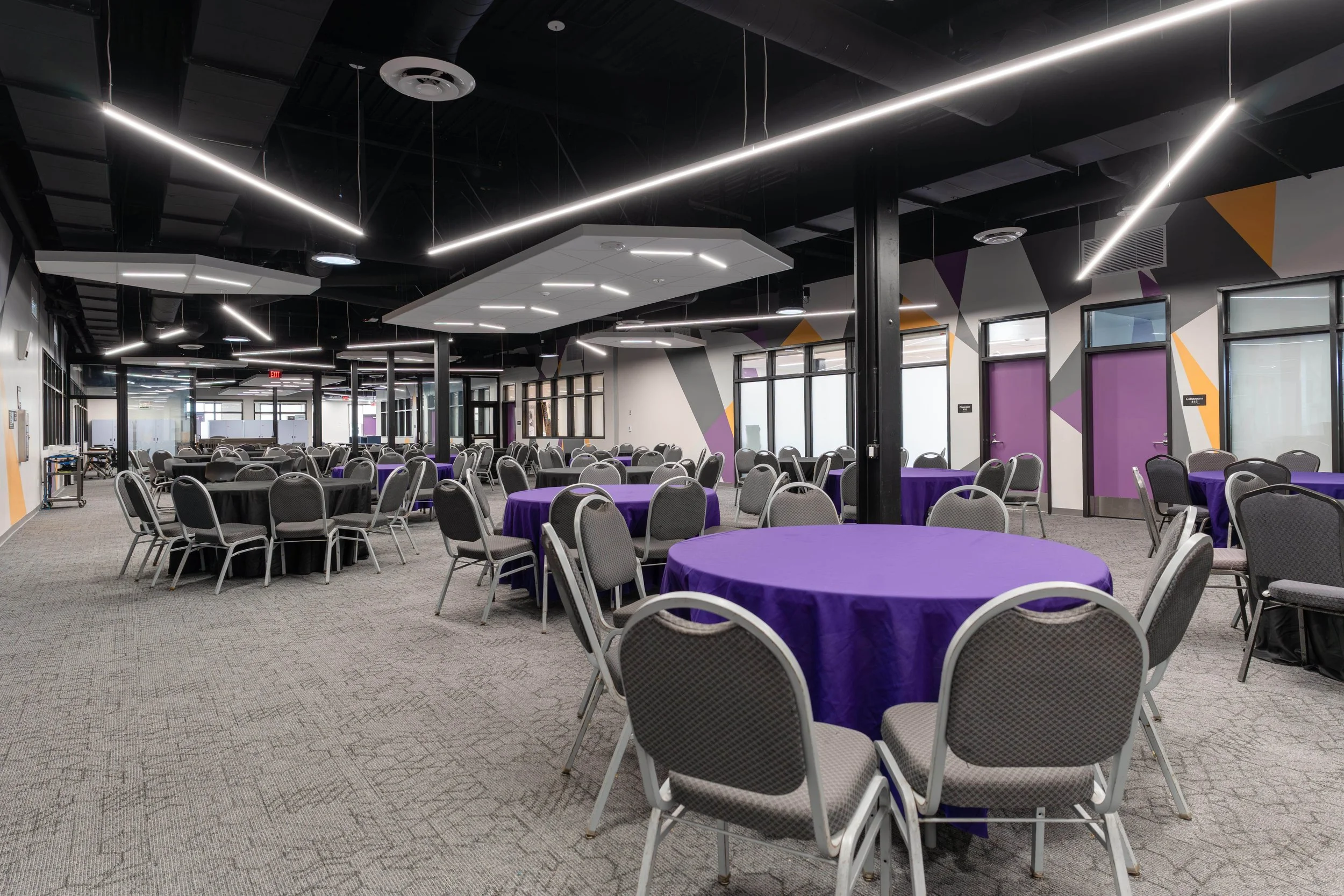 Empty banquet hall with round tables covered with purple tablecloths, gray chairs, modern ceiling lights, and colorful wall accents.