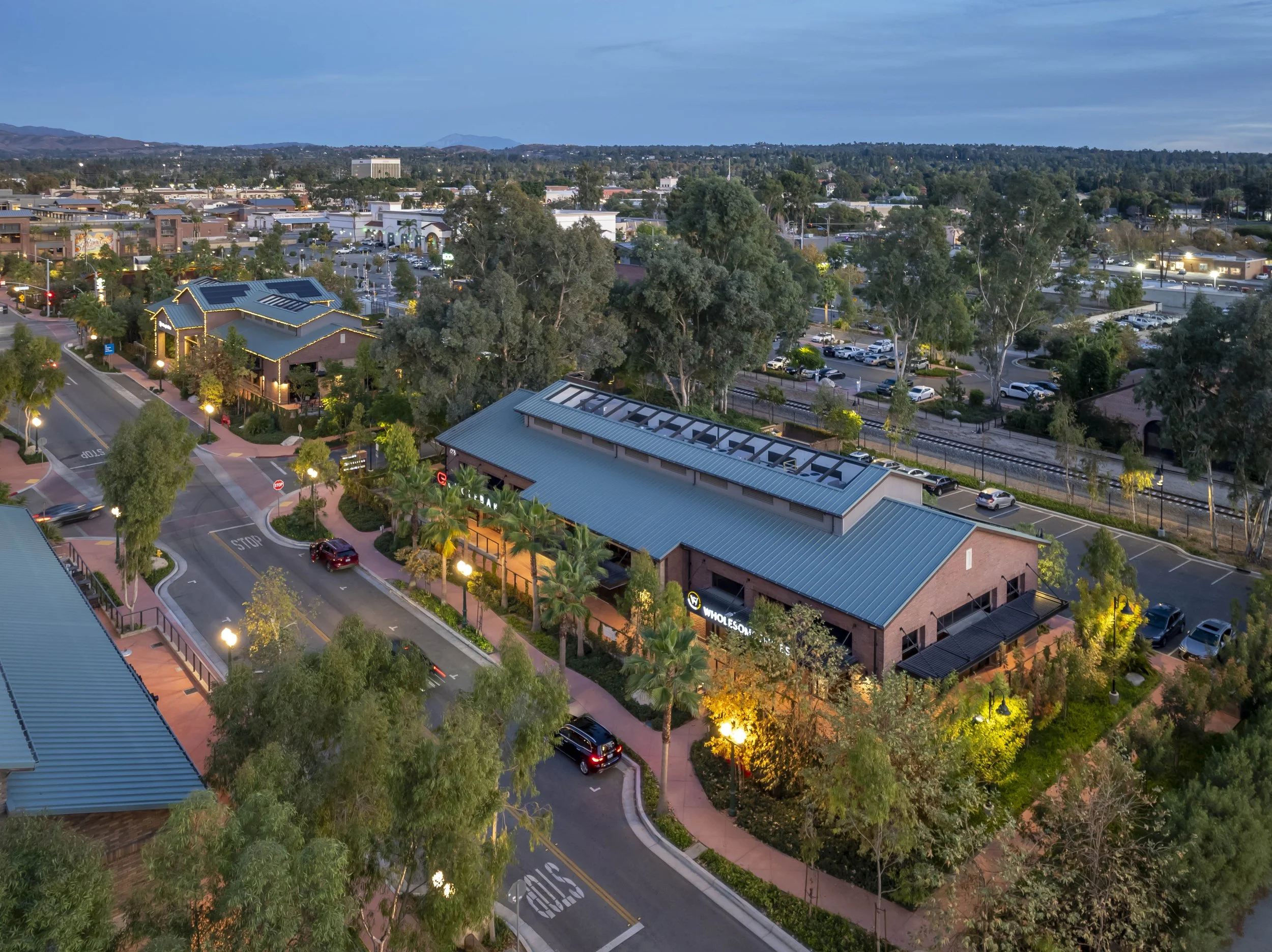 An aerial view of a shopping area at dusk, featuring a large building with a blue metal roof labeled 'Whole Foods,' surrounded by trees, parking lots, and nearby streets with cars.