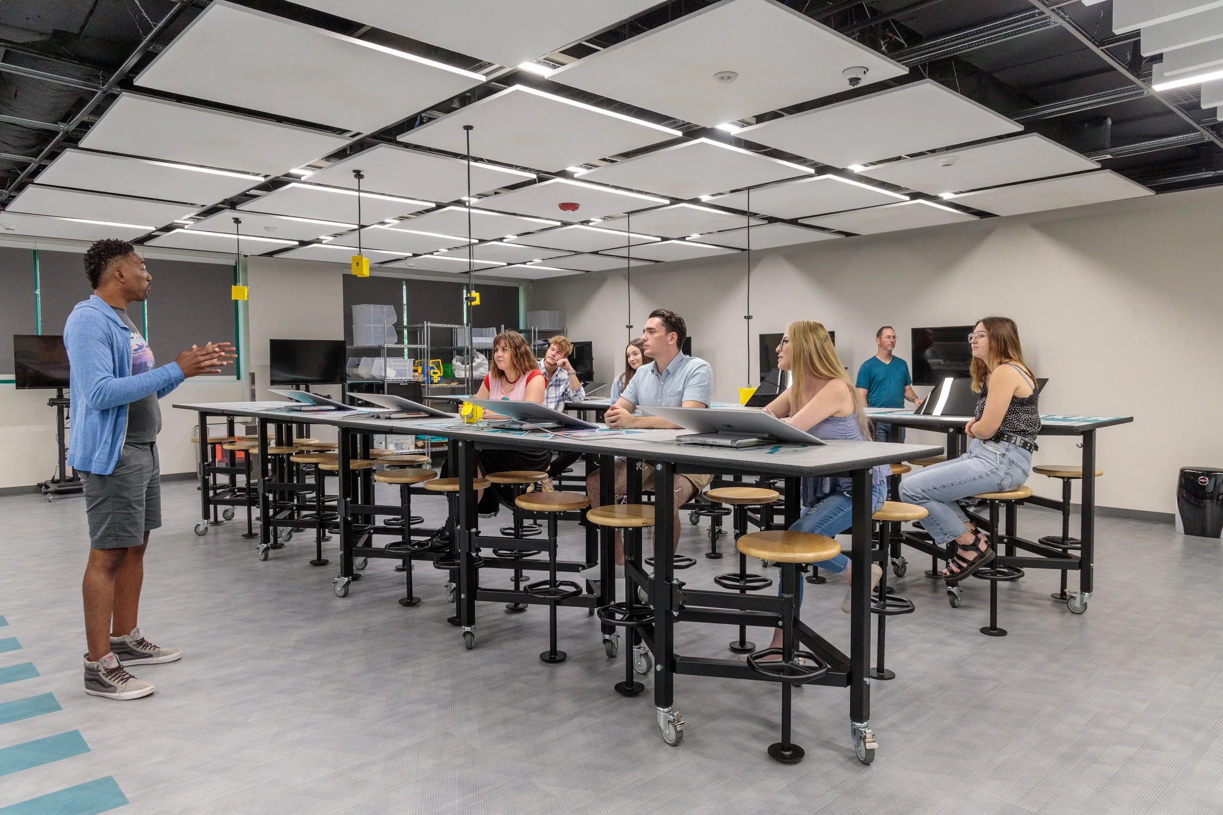 Instructor giving a lecture to students seated on high chairs in a modern classroom or laboratory with bright lighting and technology equipment.