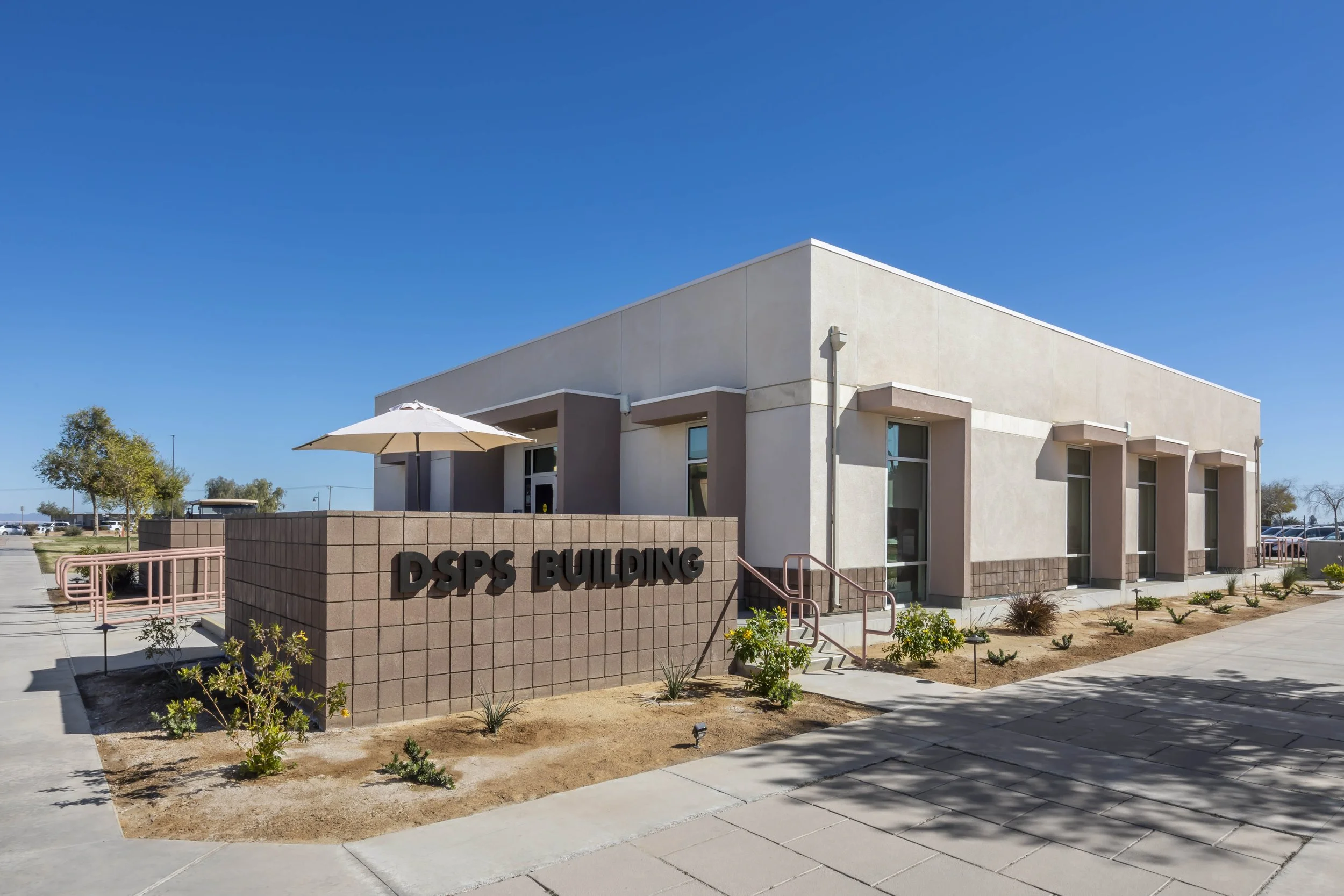 Exterior view of the DSPS building with a sign, small landscaped garden, and outdoor seating area under a white umbrella, on a sunny day with a clear blue sky.