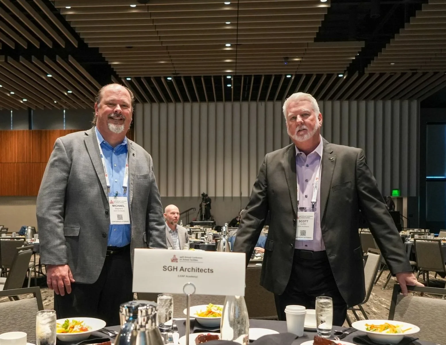 Two men in suits standing behind a conference table at an event, with a sign reading "SGH Architects" in front of them, in a modern conference hall.