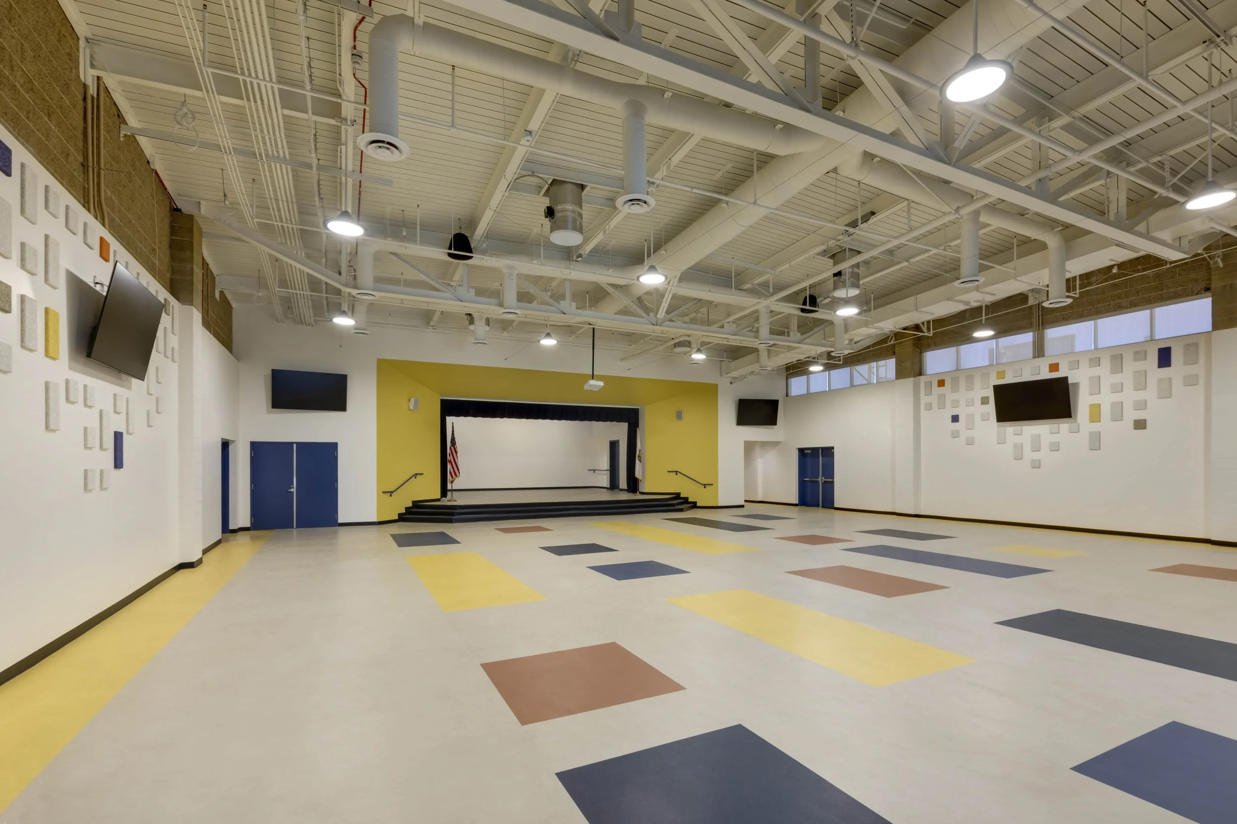 Empty school auditorium or multipurpose room with a small stage at the front, American flag, and multiple wall-mounted televisions, decorated with square wall art in various colors.