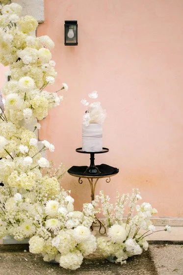 A decorated cake on a black stand, surrounded by white flowers and set against a pink wall.