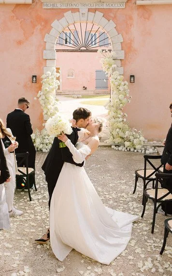 Bride and groom kissing at their wedding ceremony, with guests and an archway decorated with white flowers in the background.