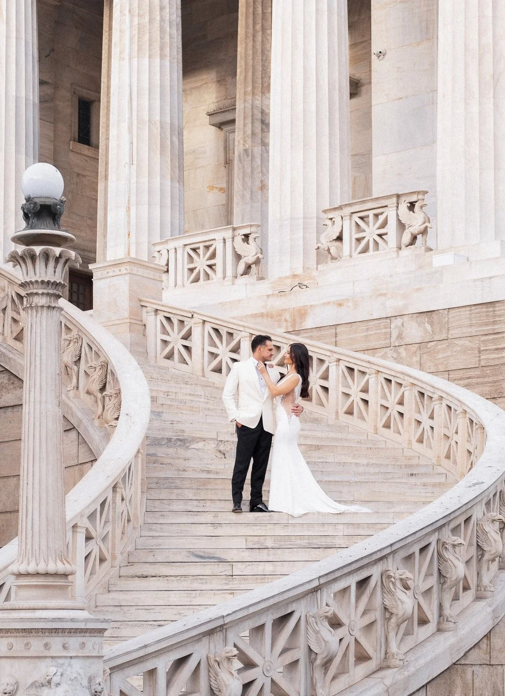A bride and groom standing on a grand marble staircase outside a historic building, gazing at each other. The bride is in a white wedding gown, and the groom is in a white blazer and black pants.
