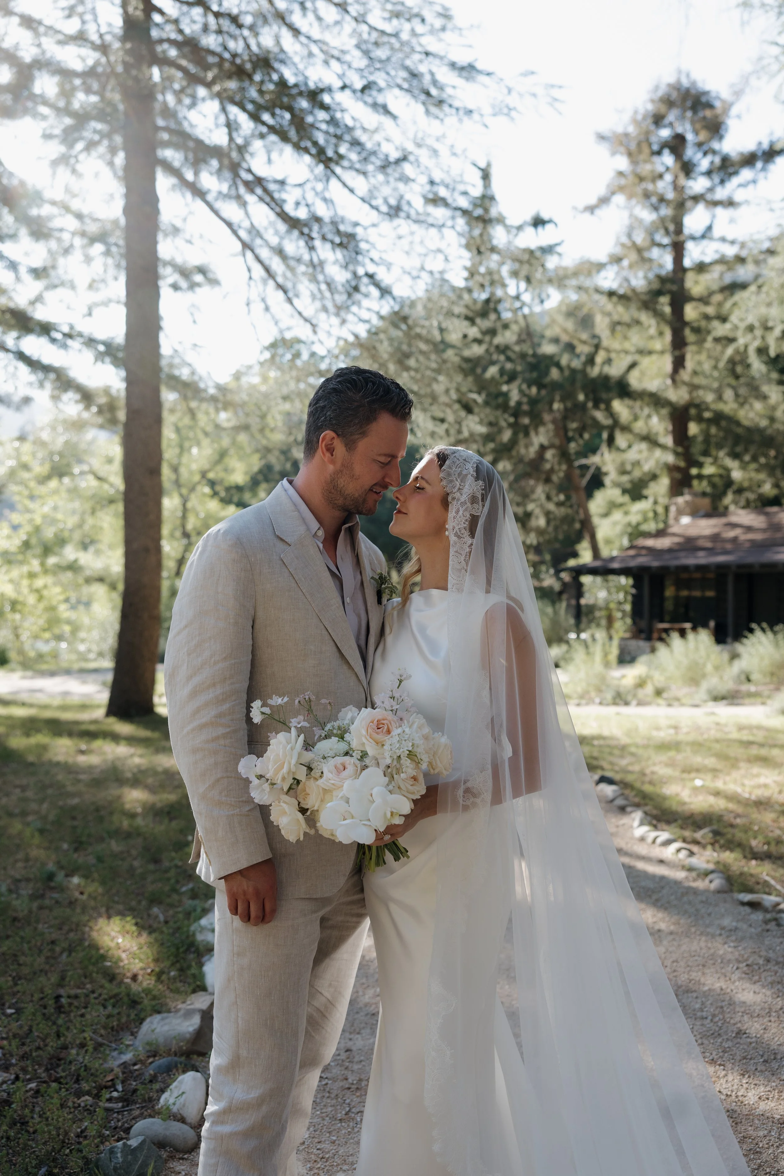 A bride and groom are standing close together outdoors in a wooded area, gazing into each other's eyes with a backdrop of trees and sunlight filtering through the leaves. The bride is wearing a white wedding dress with a lace veil and holding a bouqu