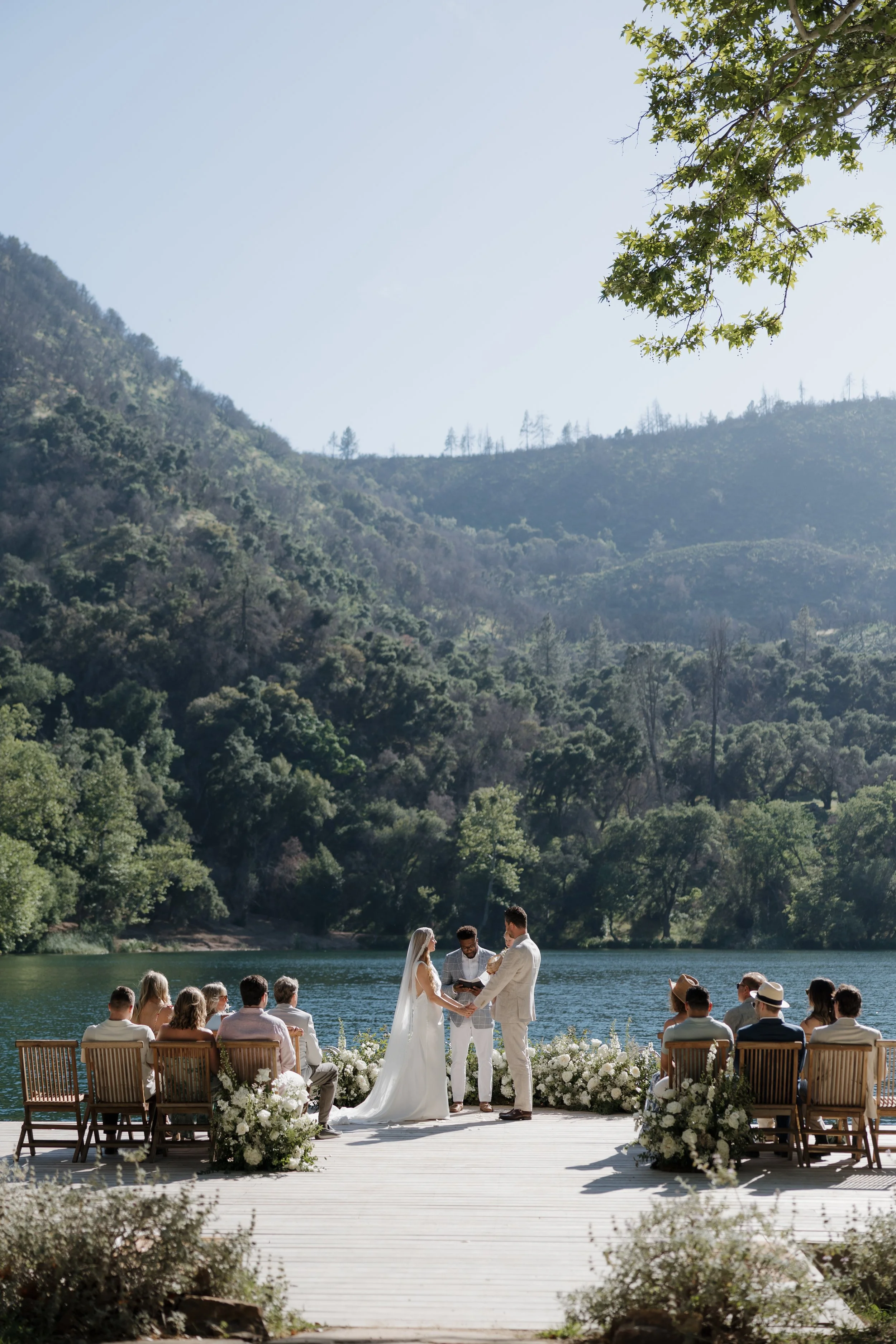 A wedding ceremony taking place outdoors on a lakeside deck with mountains and trees in the background, guests seated on chairs, with the bride and groom exchanging vows.