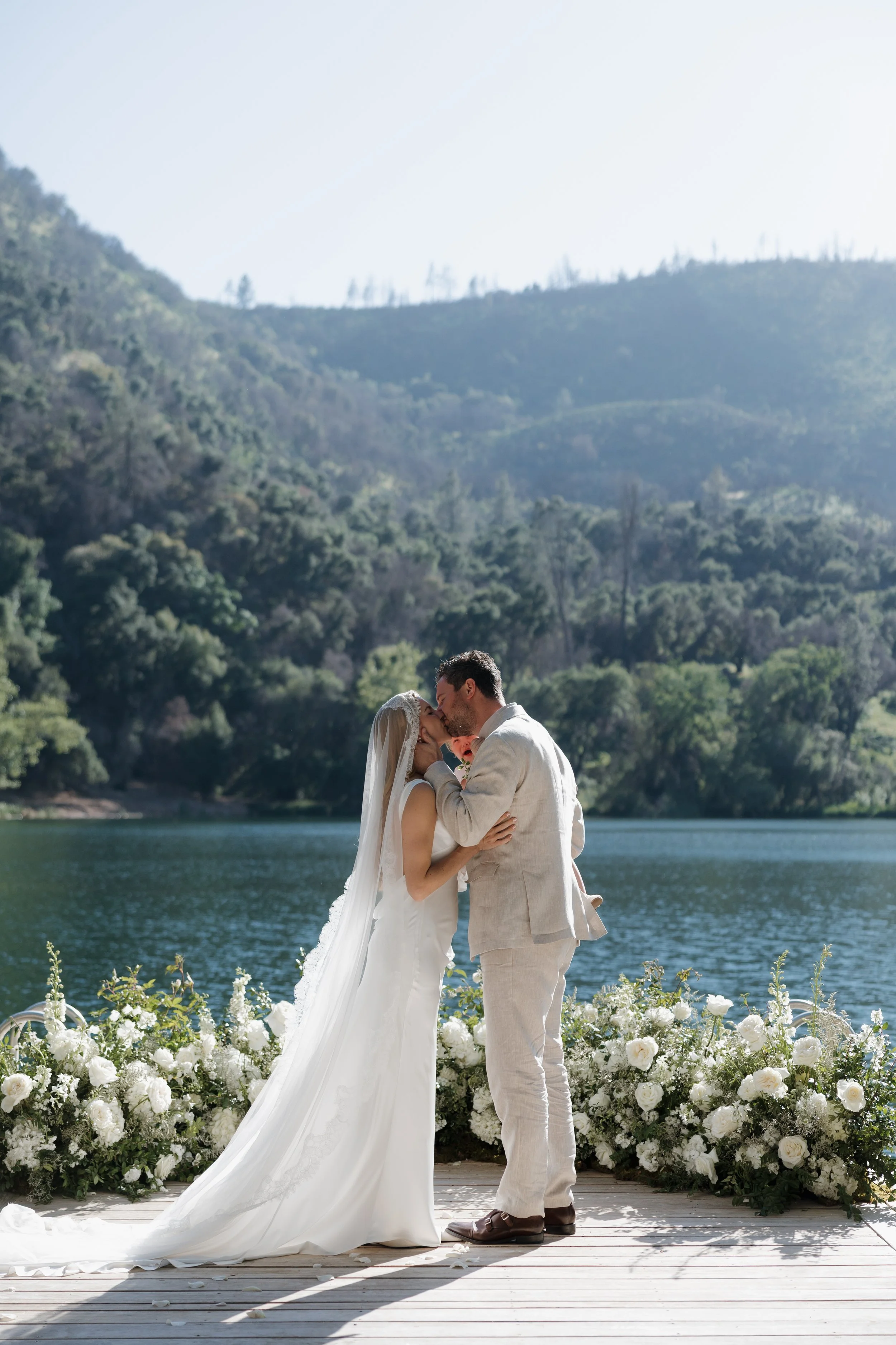 A bride and groom sharing a kiss at their wedding ceremony outdoors by a lake, surrounded by white flowers and green trees with mountains in the background.