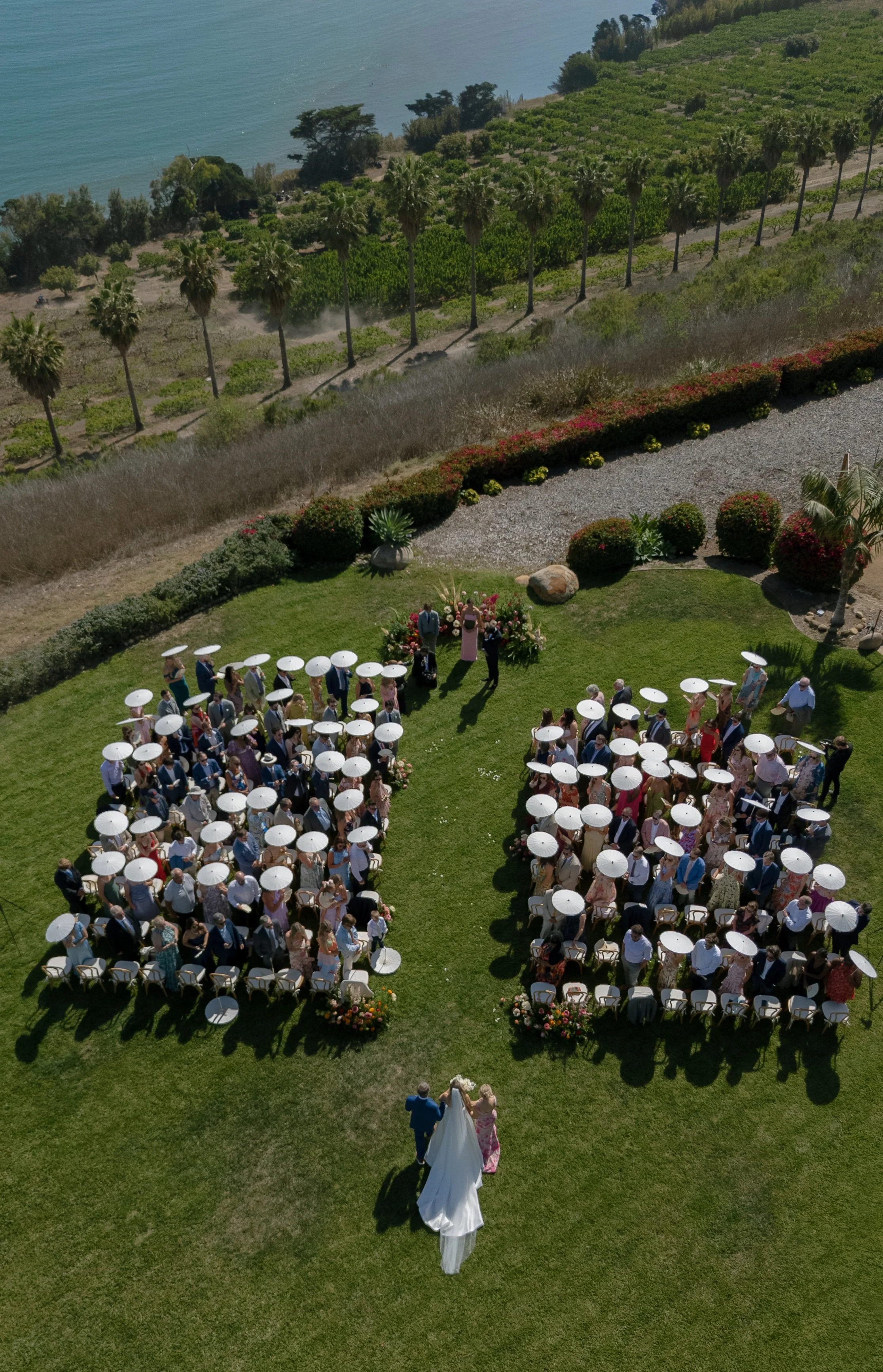 An outdoor wedding ceremony taking place on a grassy lawn overlooking the ocean. Guests seated under white umbrellas watch as a bride and groom stand in front of a floral arrangement, with a person in pink officiating.