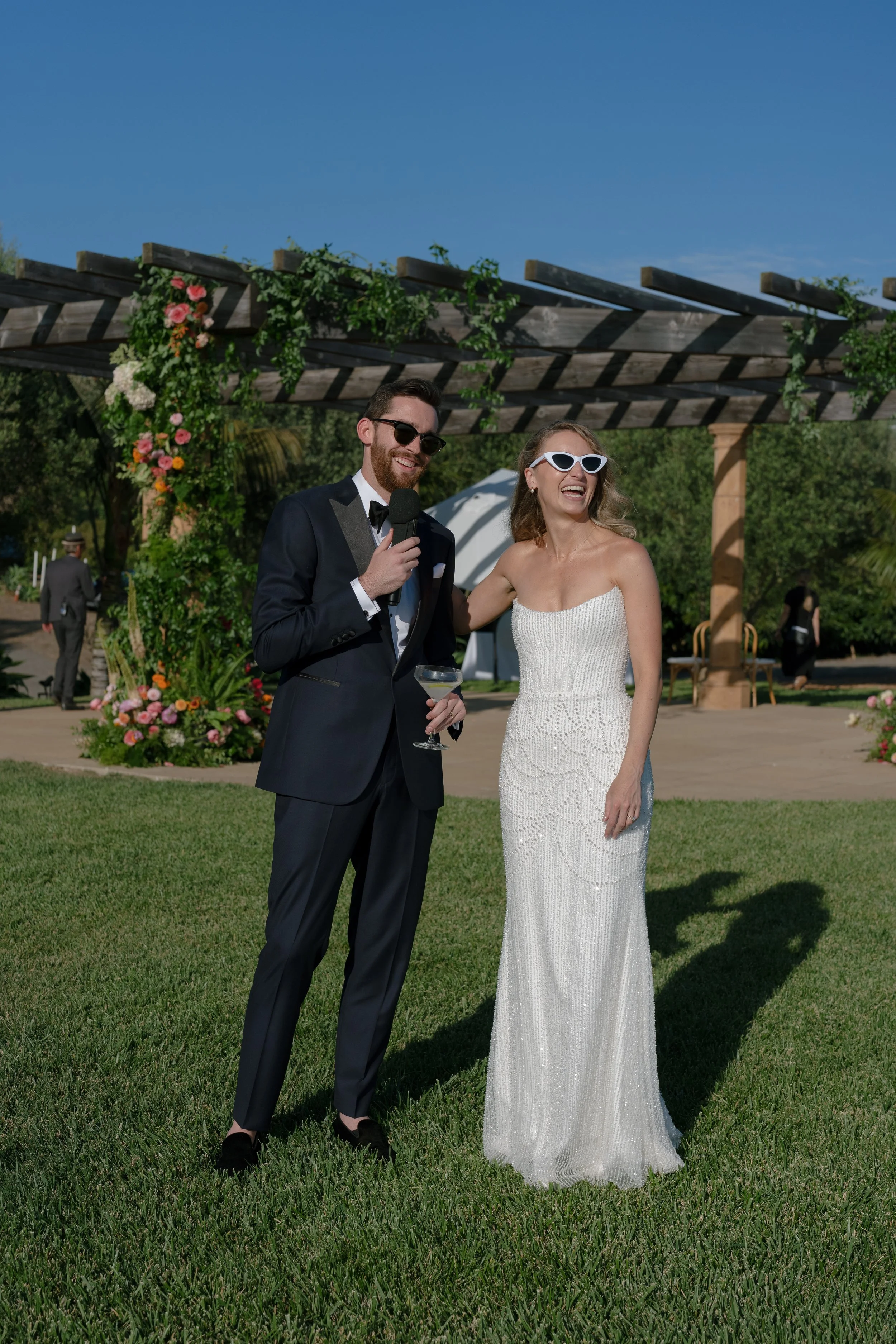 A man and a woman outdoors at a wedding, smiling, with a floral arch and greenery in the background. The man is wearing a tuxedo and sunglasses, holding a microphone and a drink, and the woman is wearing a white strapless gown and sunglasses.