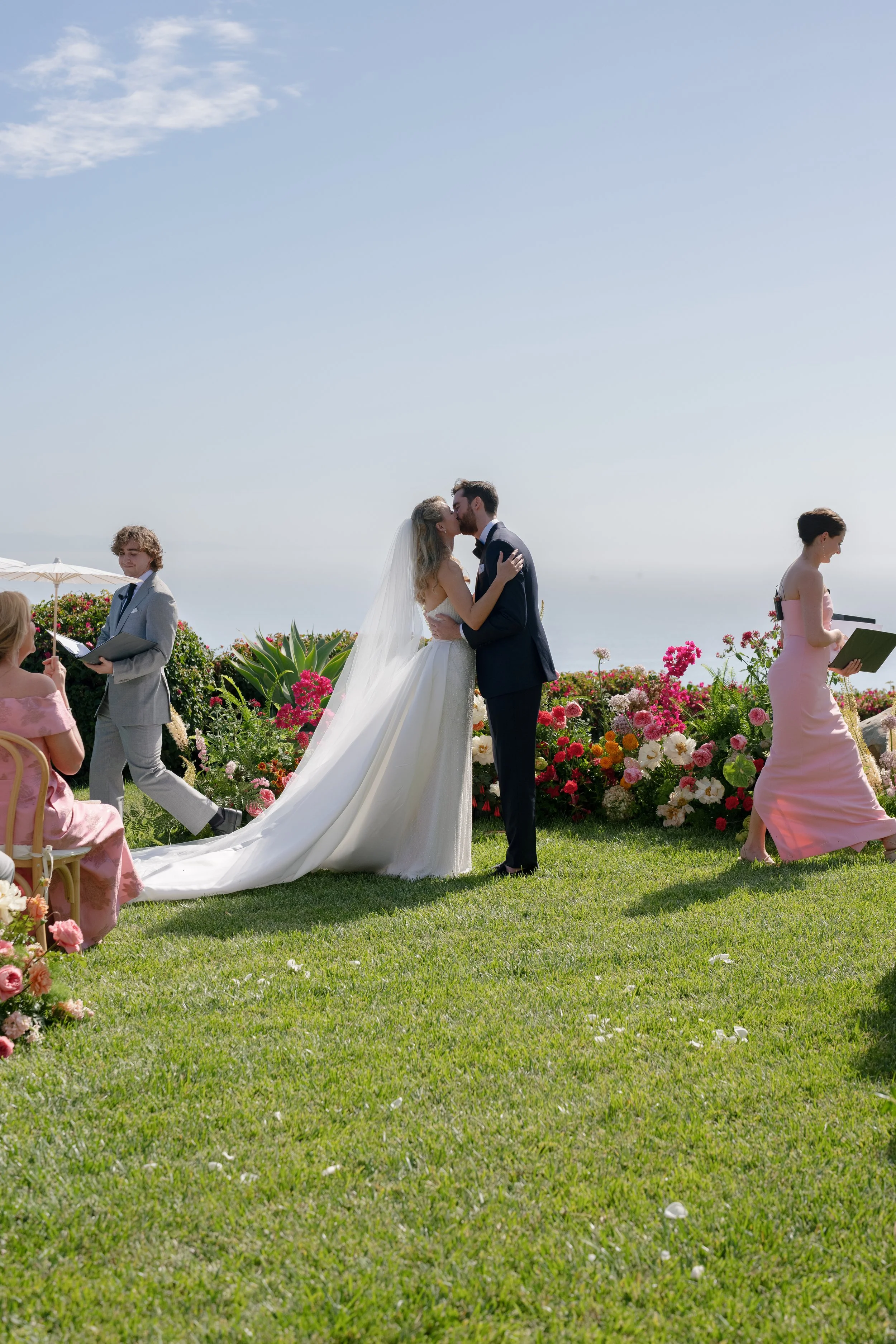 A wedding ceremony taking place outdoors with a bride and groom kissing, surrounded by flowers and guests.