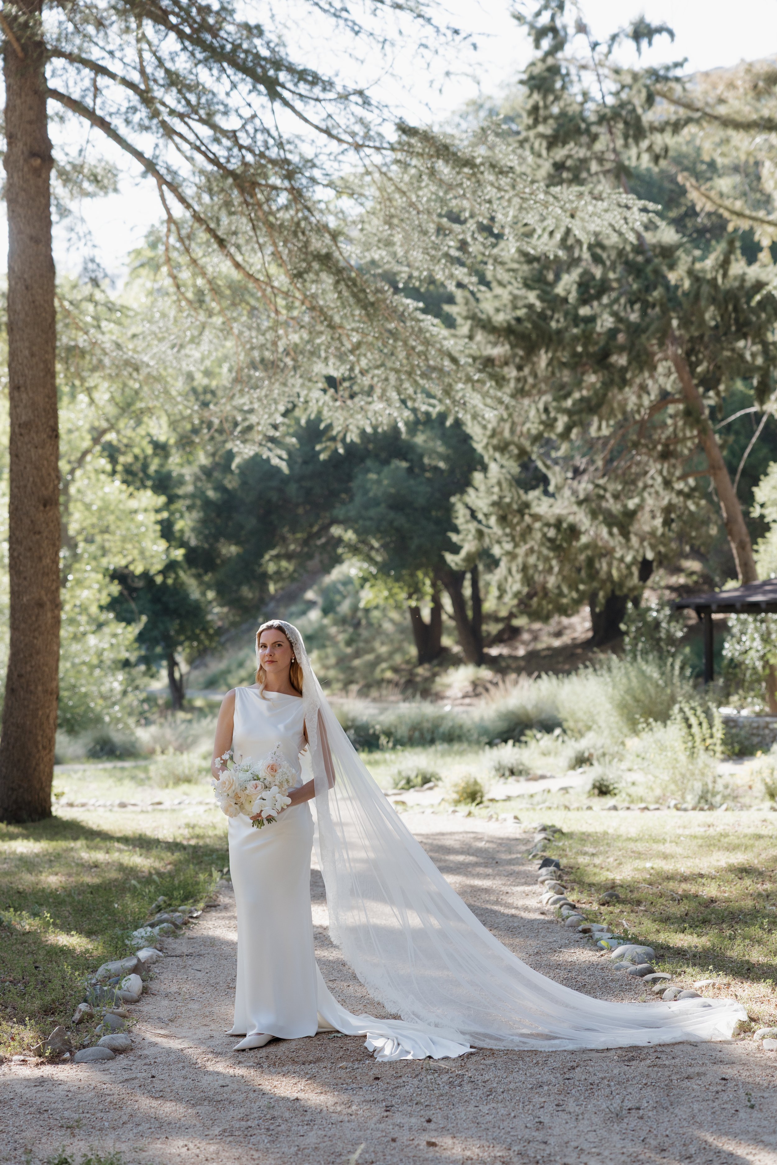 A bride in a white wedding dress with a veil, holding a bouquet, standing on a garden path surrounded by trees and greenery, sunlight filtering through the leaves.