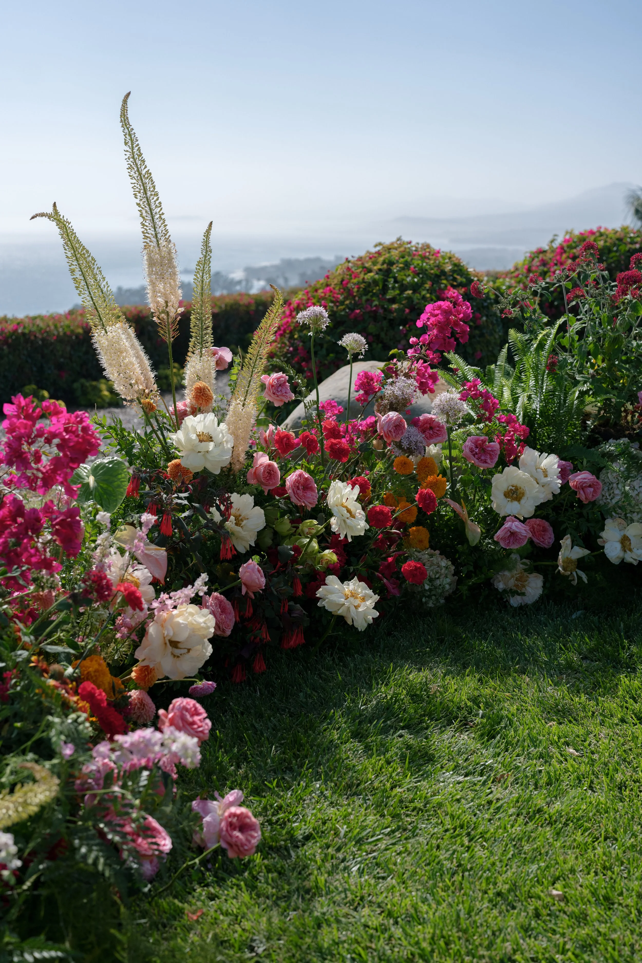 Colorful garden with pink, white, and red flowers, green grass, and a view of hills and water in the background on a sunny day.