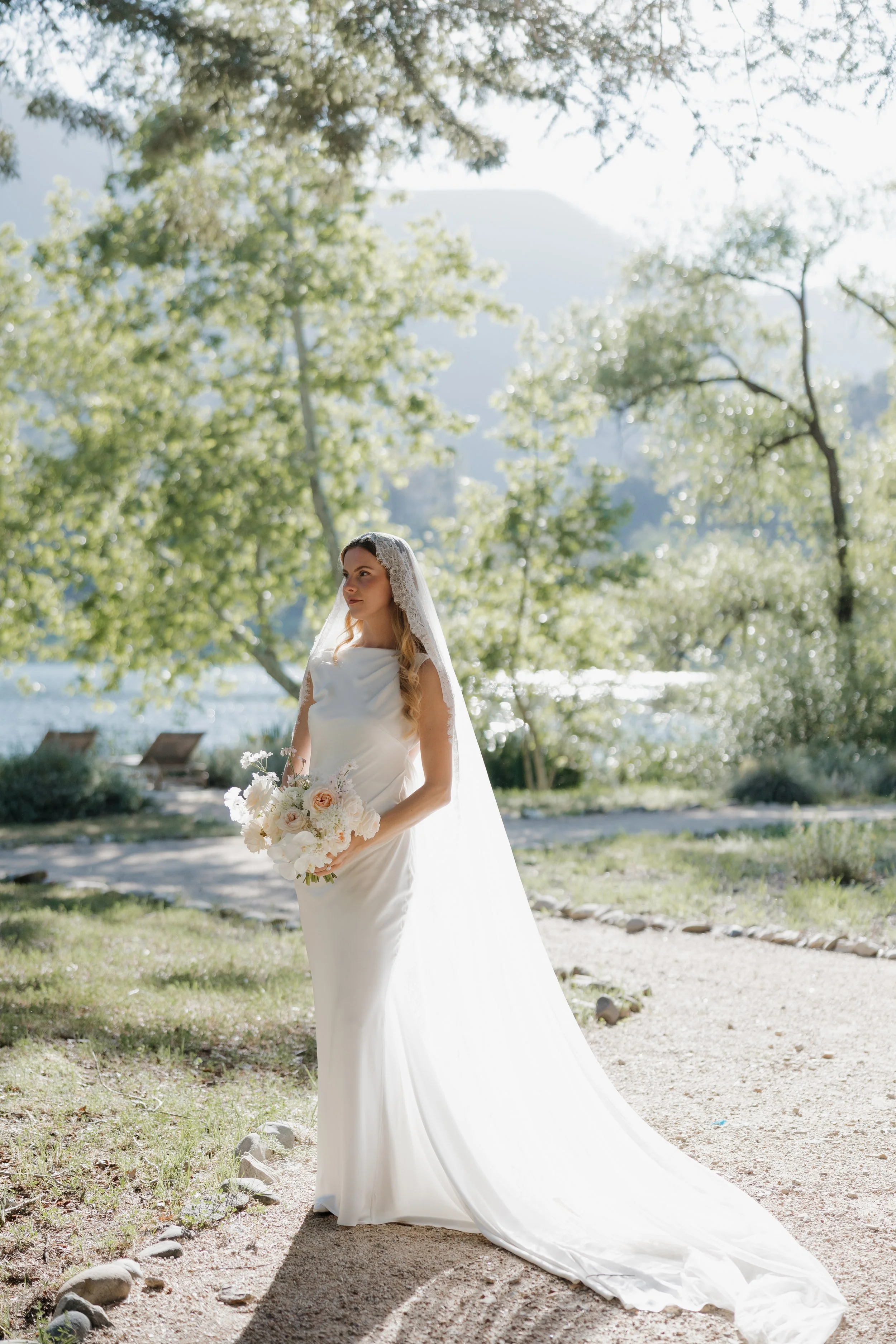 A bride in a white wedding gown holding a bouquet of white and light pink flowers, standing outdoors on a dirt path surrounded by trees and greenery, with sunlight filtering through the leaves.