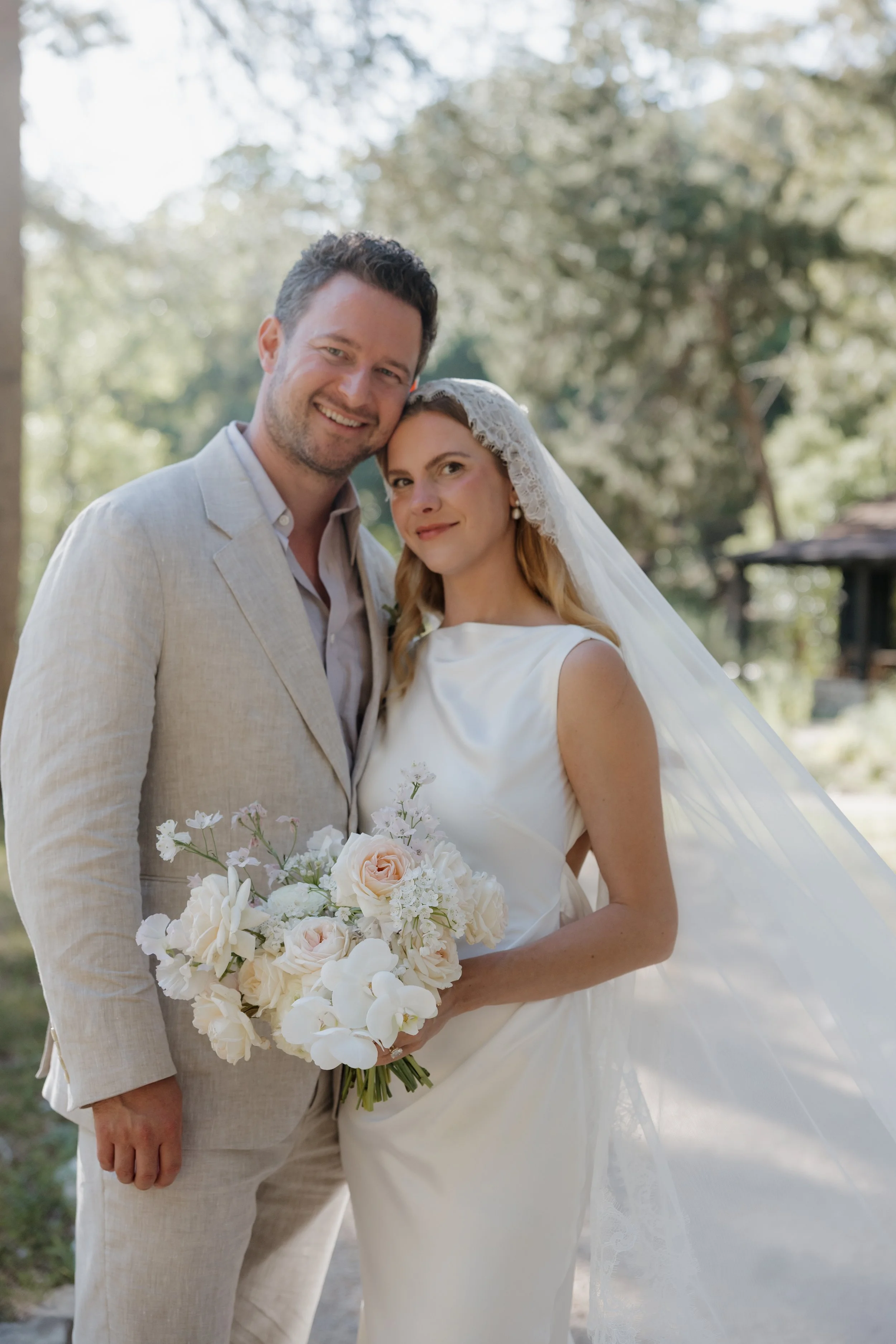A bride and groom smiling outdoors on their wedding day, with the bride holding a bouquet of white and blush flowers, dressed in a satin wedding gown with lace veil, and the groom wearing a light-colored suit with an open collar shirt.