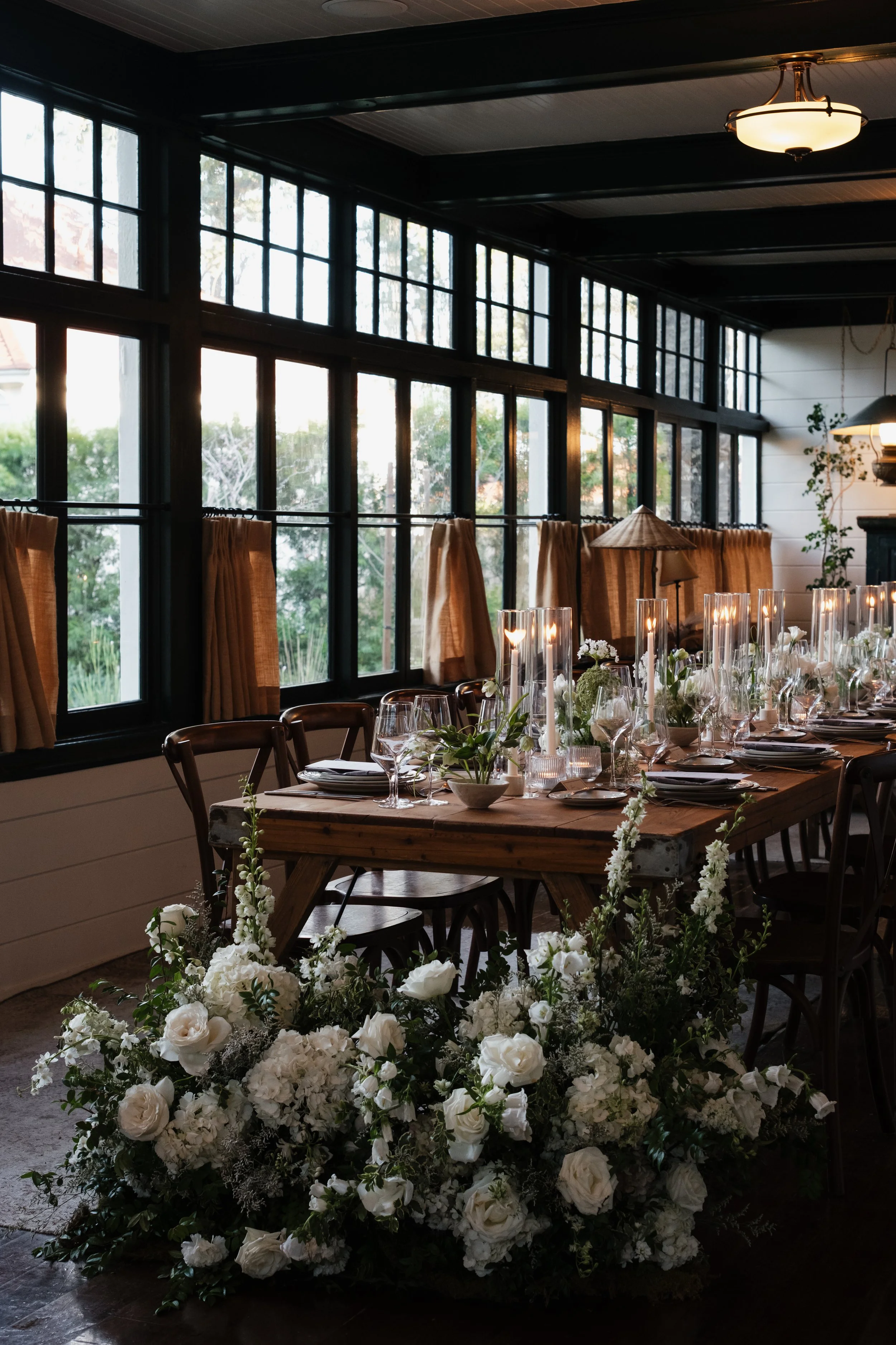 Elegant indoor dining setup with a wooden table and white floral centerpiece, surrounded by lined-up chairs, illuminated by candlelit glasses and warm ambient lighting, with large windows and curtains in the background.