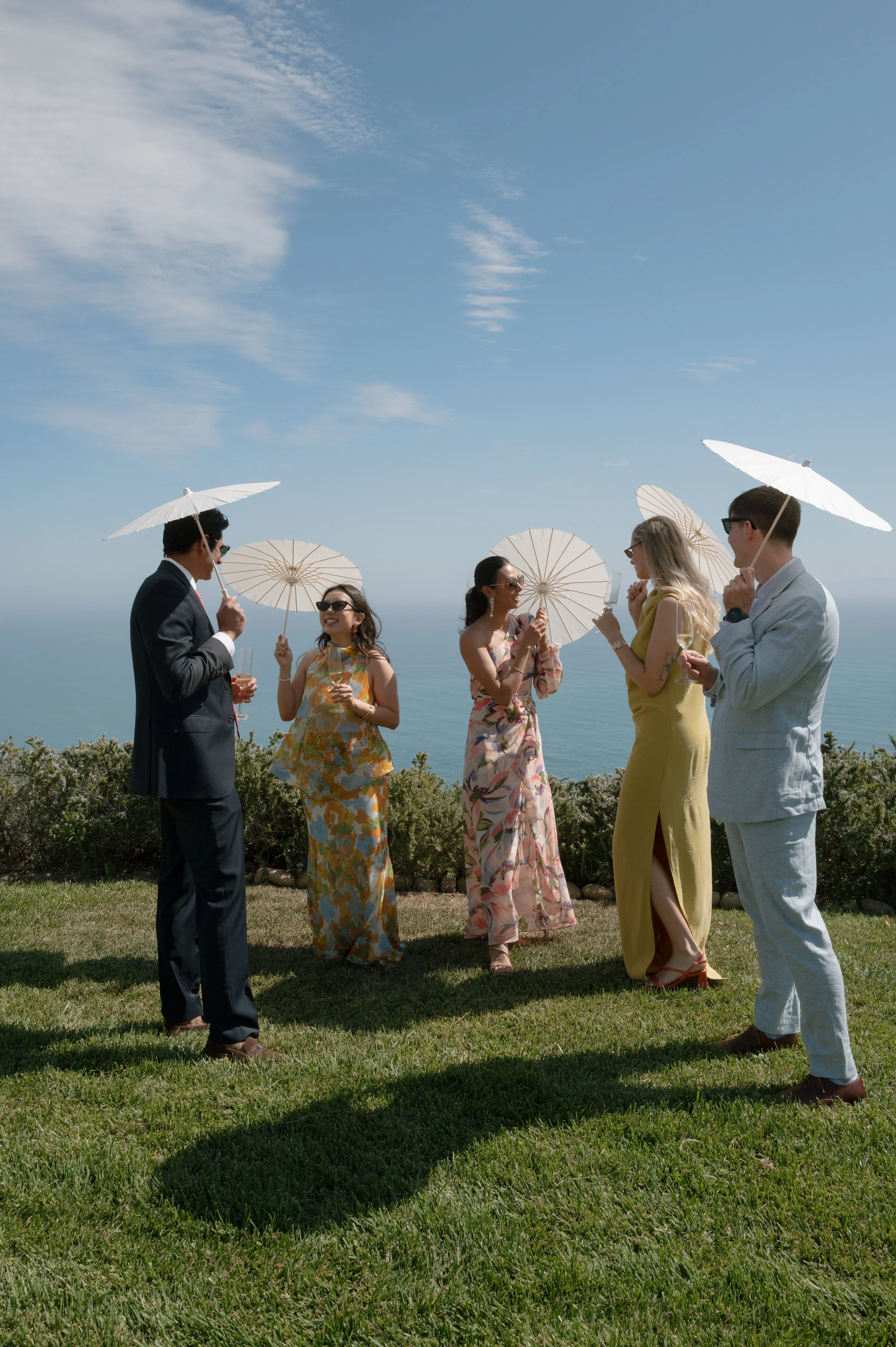 A group of six people at an outdoor gathering, holding umbrellas, wearing formal and colorful attire, with a clear sky and ocean in the background.