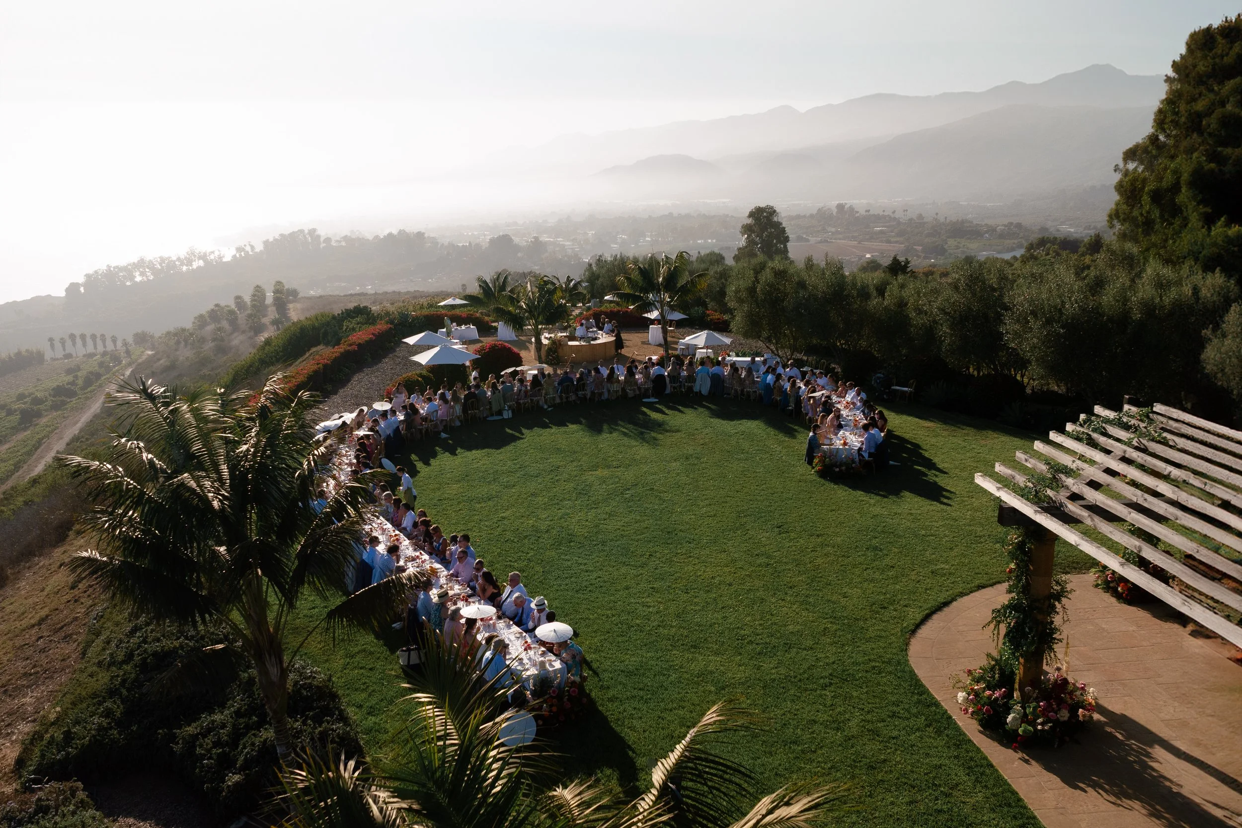 Outdoor wedding reception on a hillside with long table, white umbrellas, and guests, overlooking mountains and lush greenery.