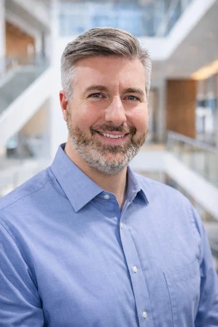 Professional man with gray hair and beard smiling, wearing a blue collared shirt, in a modern office building with glass and wooden interior.